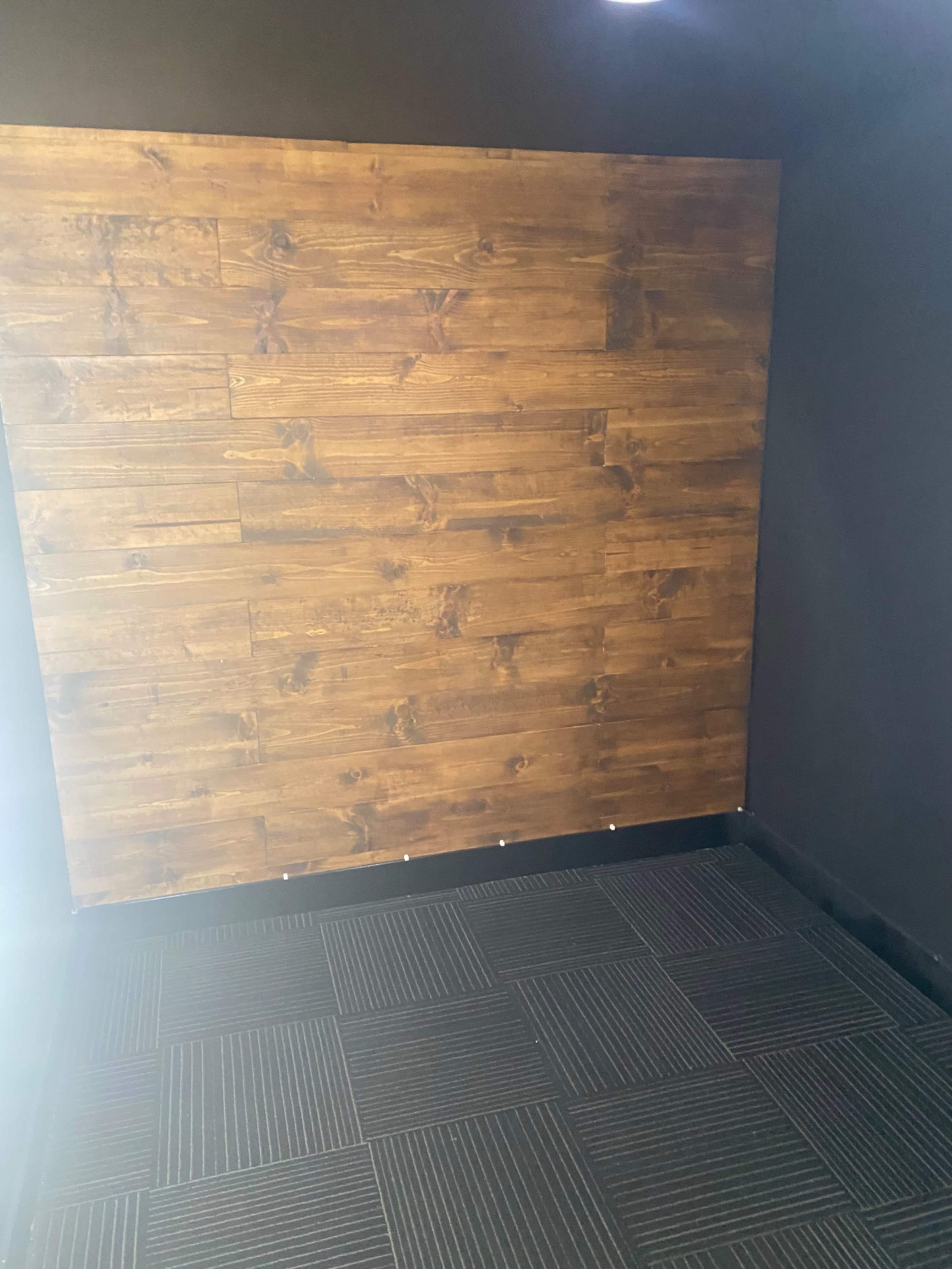 The image shows a corner of a room featuring a wooden wall with planks and a black textured floor.