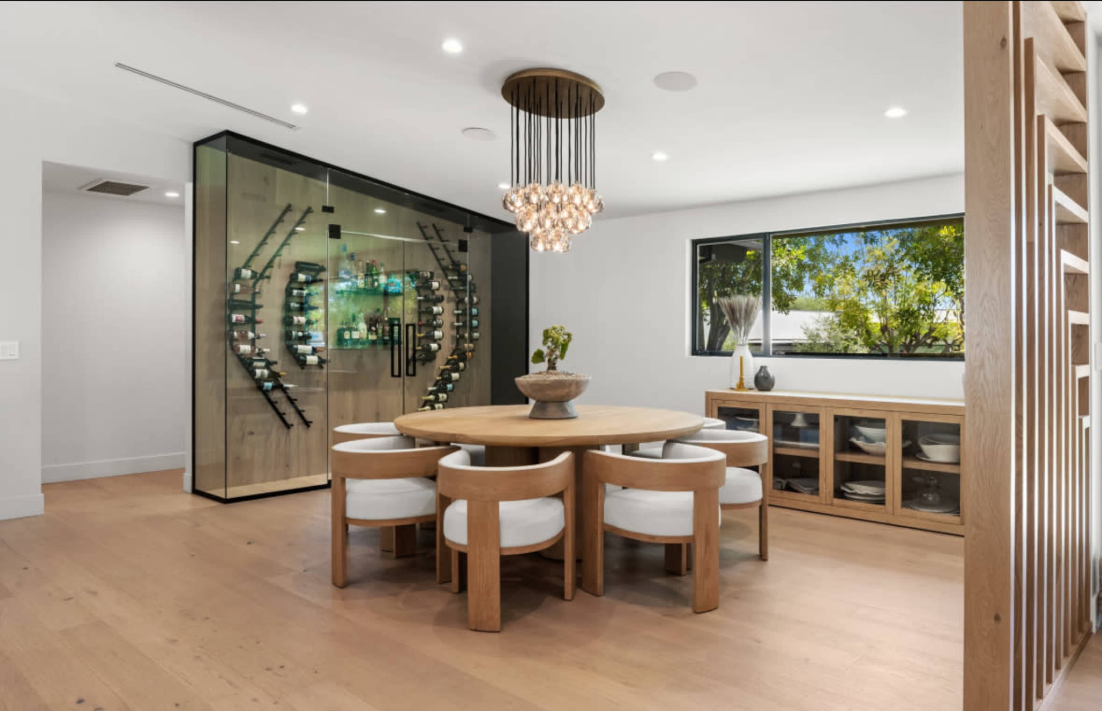 A modern dining area features a circular wooden table surrounded by white cushioned chairs, with a large window and a wine display cabinet in the background.