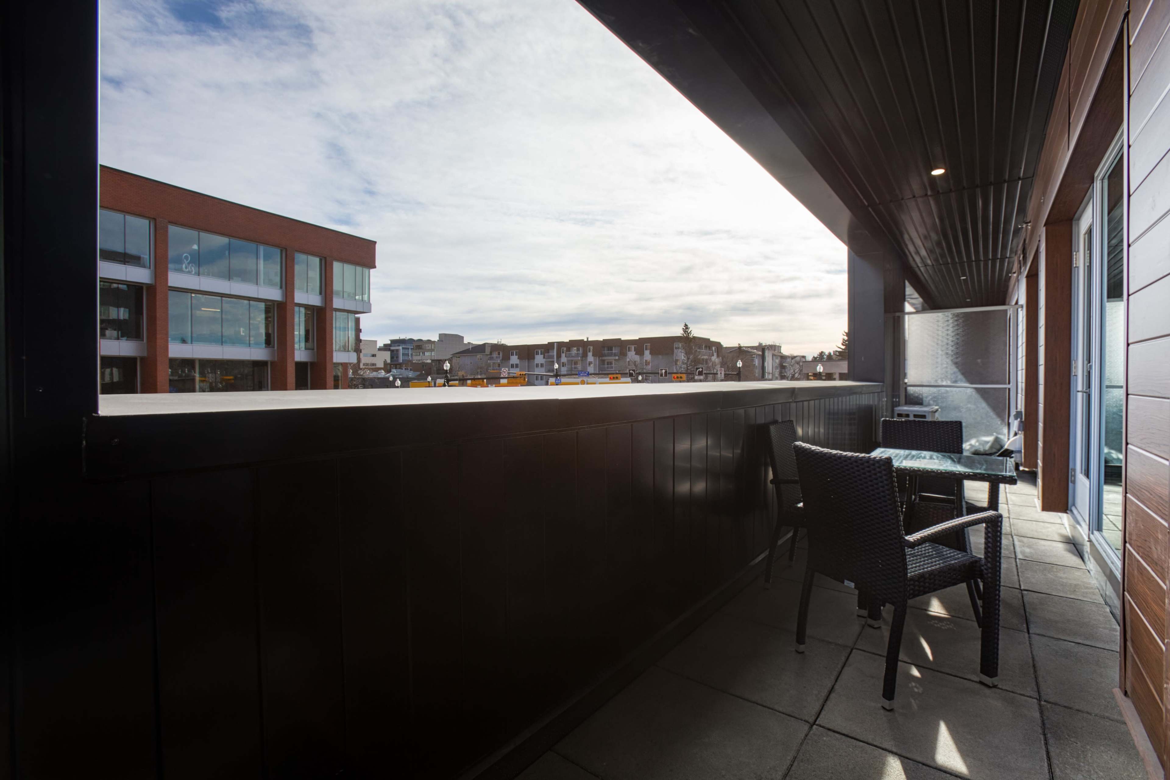 The image shows a balcony with a table and chair overlooking a cityscape under a partly cloudy sky.