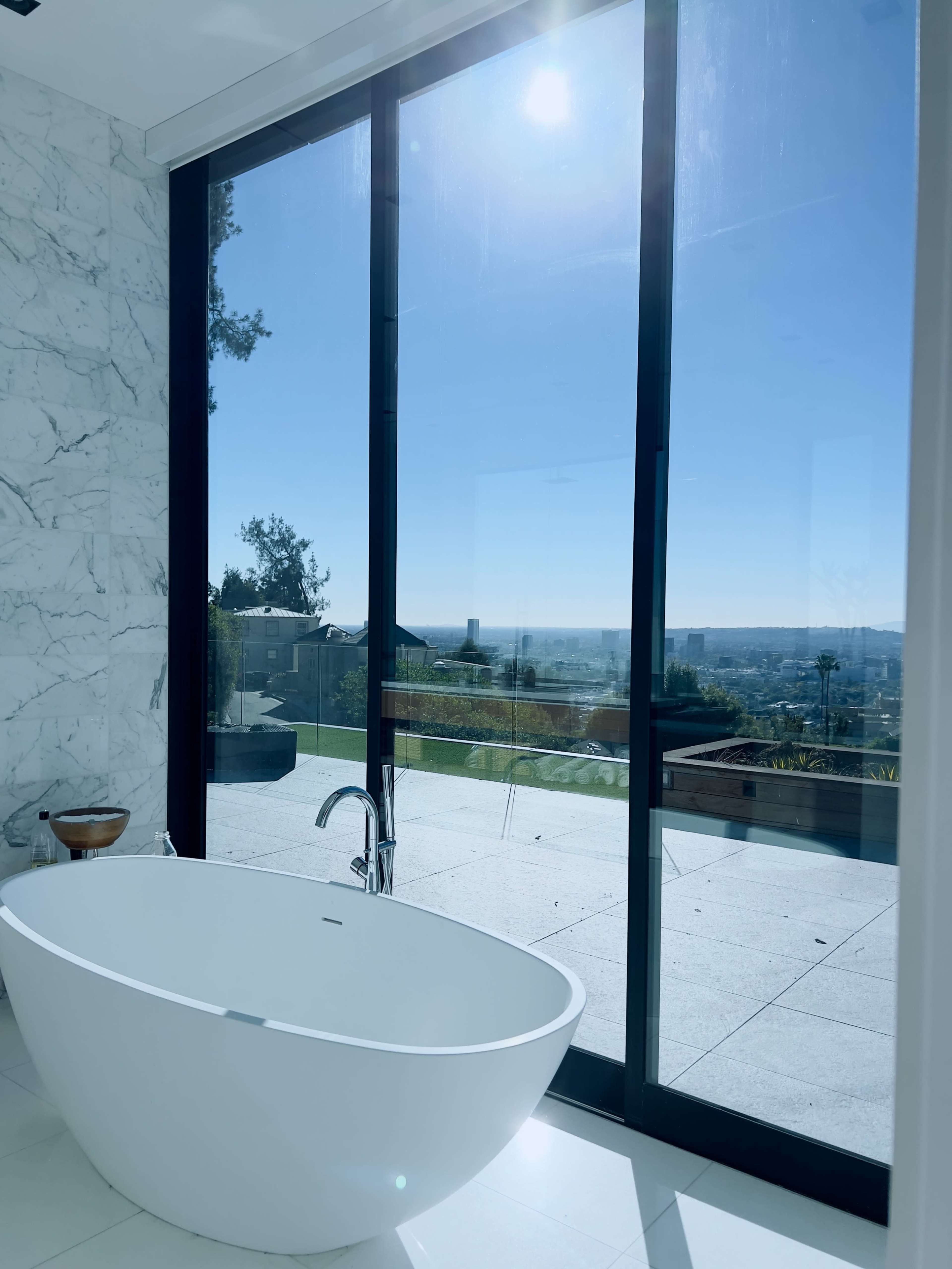 A freestanding bathtub is positioned near large glass doors that overlook a scenic landscape under a clear blue sky.