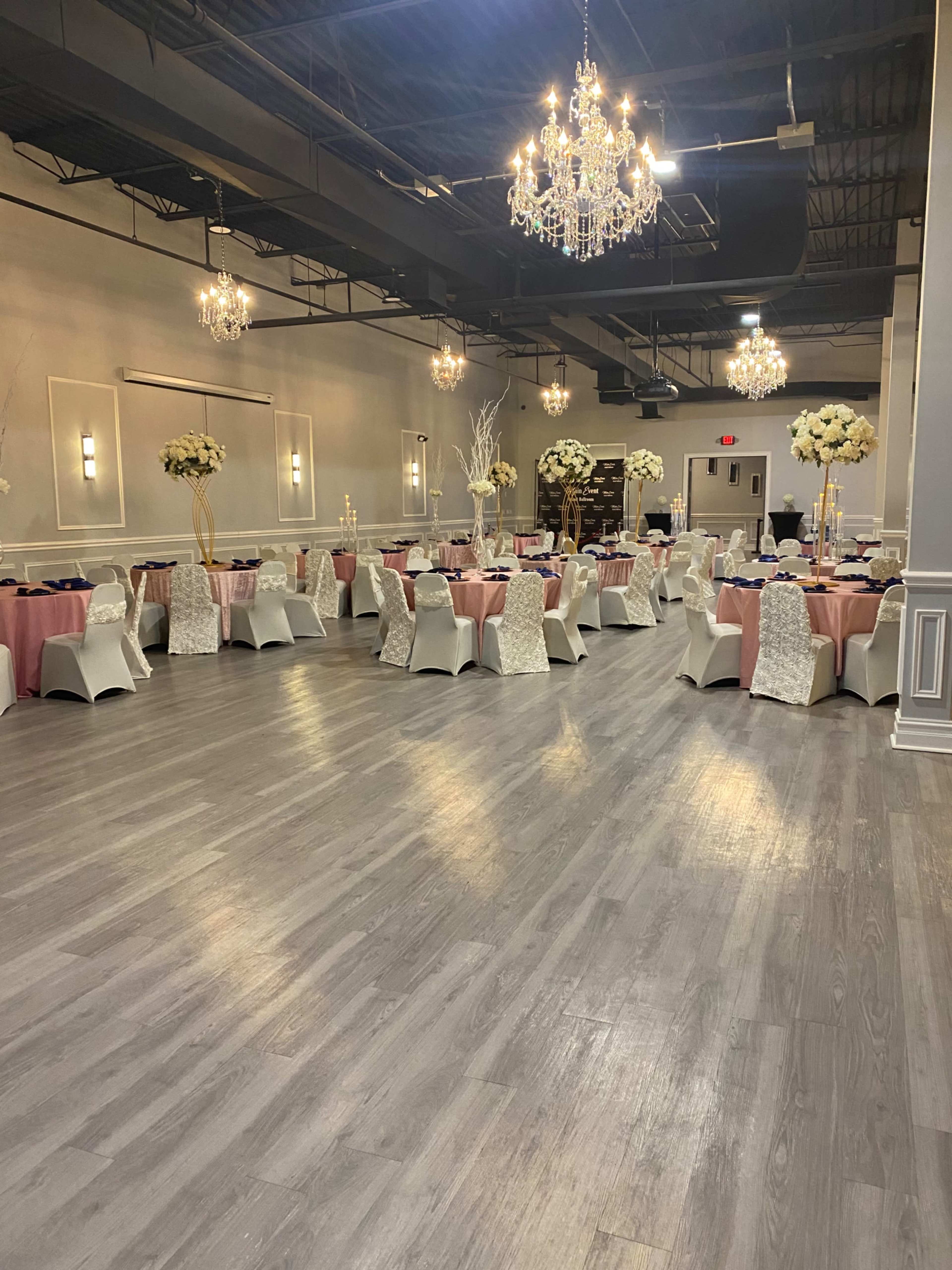 A banquet hall set up with tables covered in pink and white linens, adorned with floral centerpieces and chandeliers hanging from the ceiling.