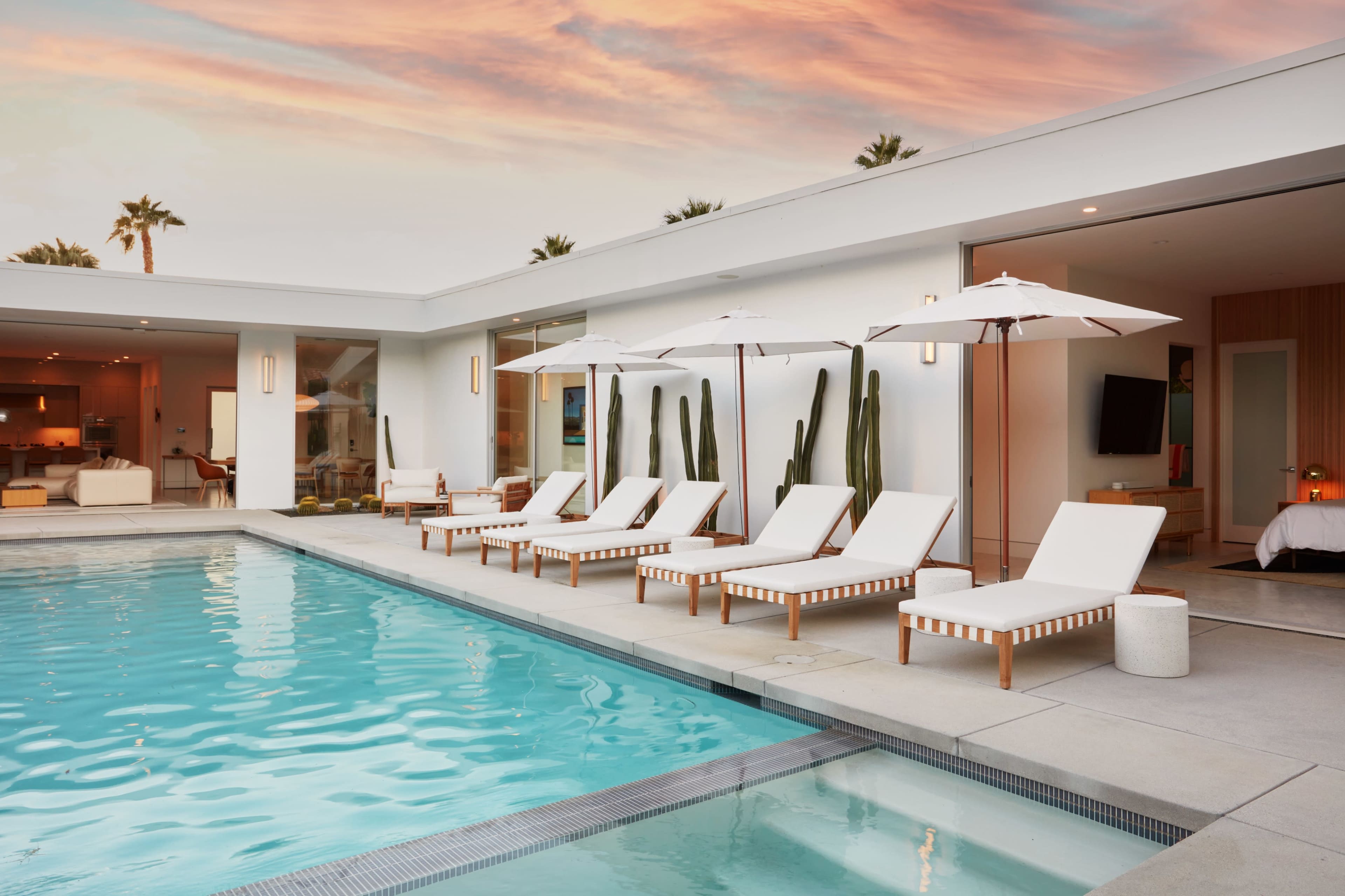 The image shows a modern pool area with lounge chairs and umbrellas beside a clear blue pool, set against a backdrop of a contemporary house and palm trees.