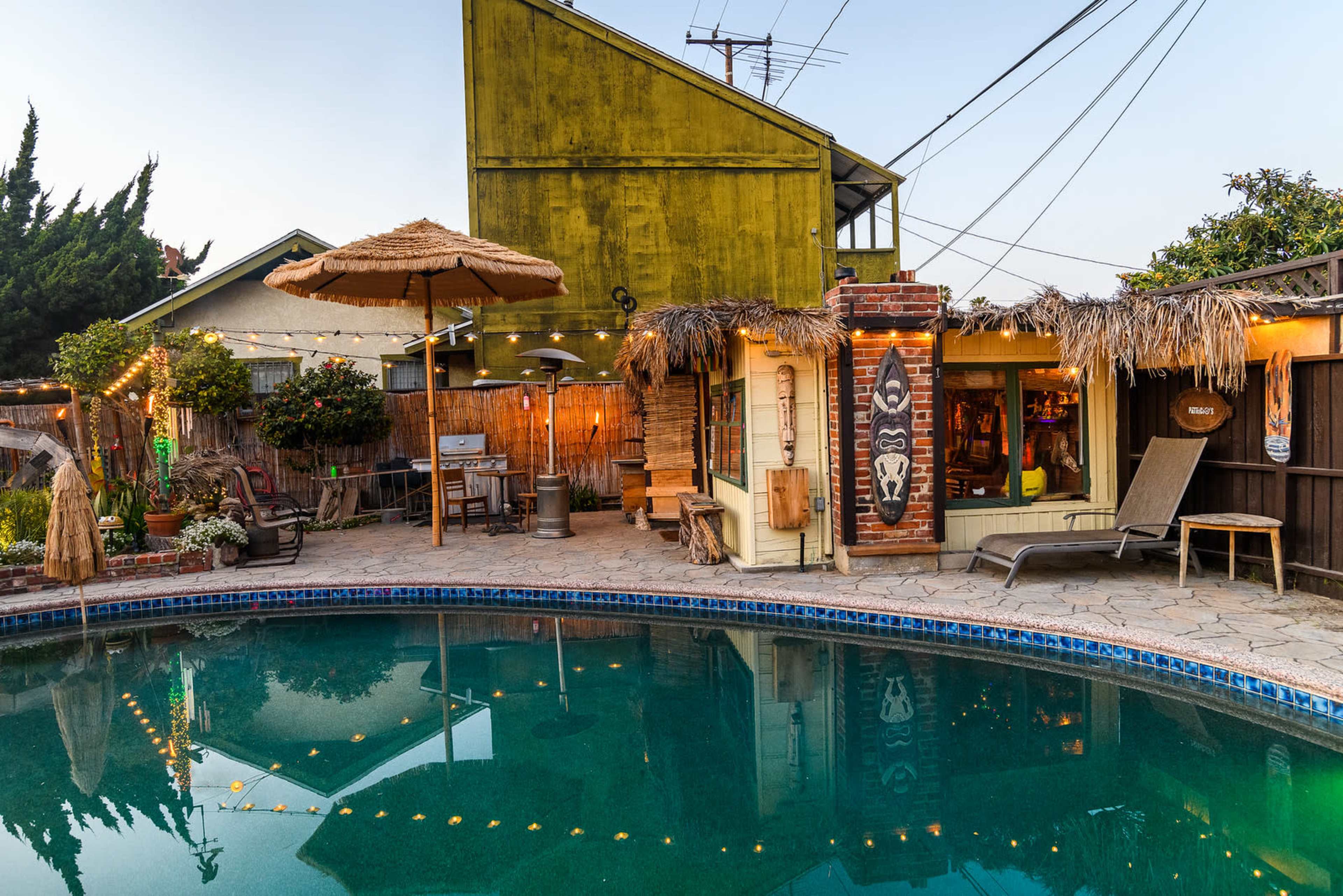 A backyard pool surrounded by a wooden deck, a tiki hut, and a two-story house with a green exterior, illuminated by string lights at dusk.