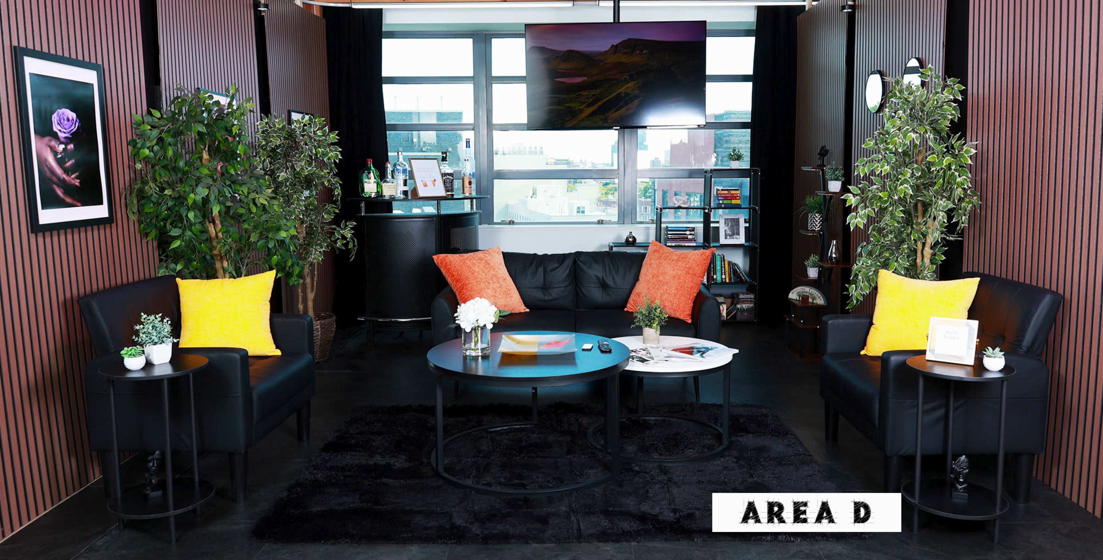 A modern lounge area features a black sofa, two armchairs, and round tables, surrounded by plants and large windows.