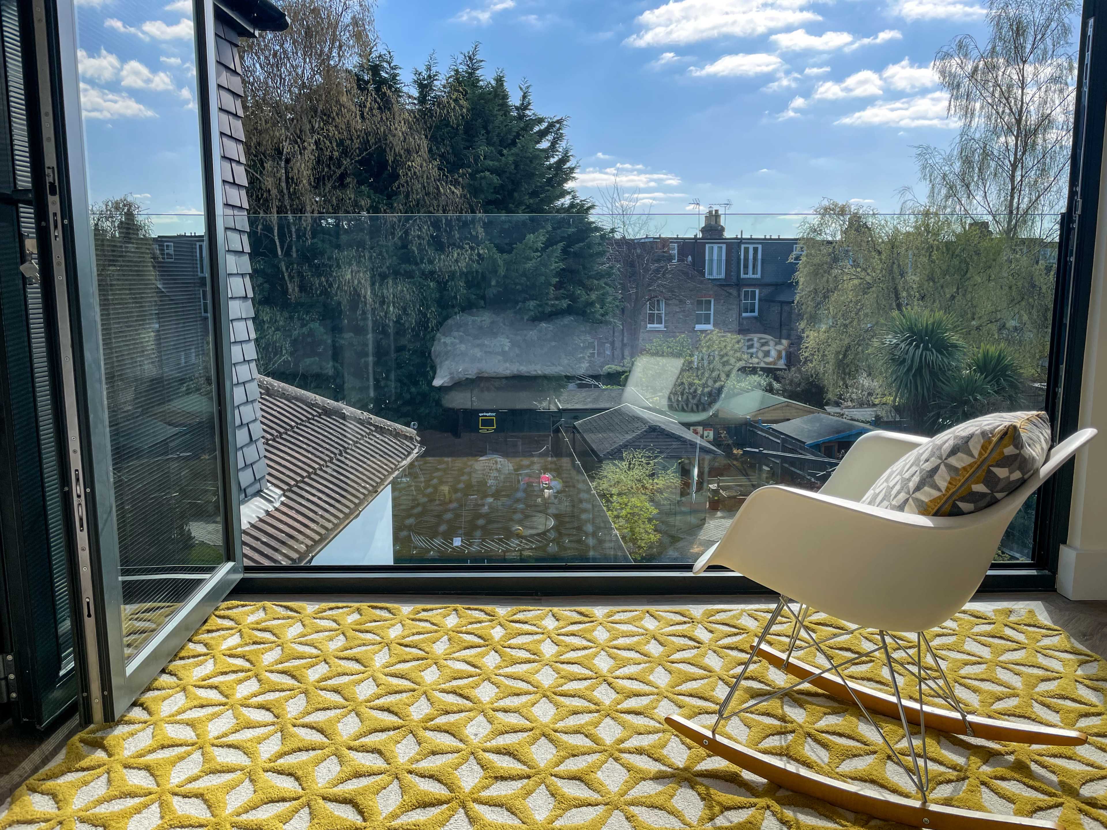 A modern interior space features a rocking chair and a geometric yellow rug, overlooking a garden and trees through large glass doors.