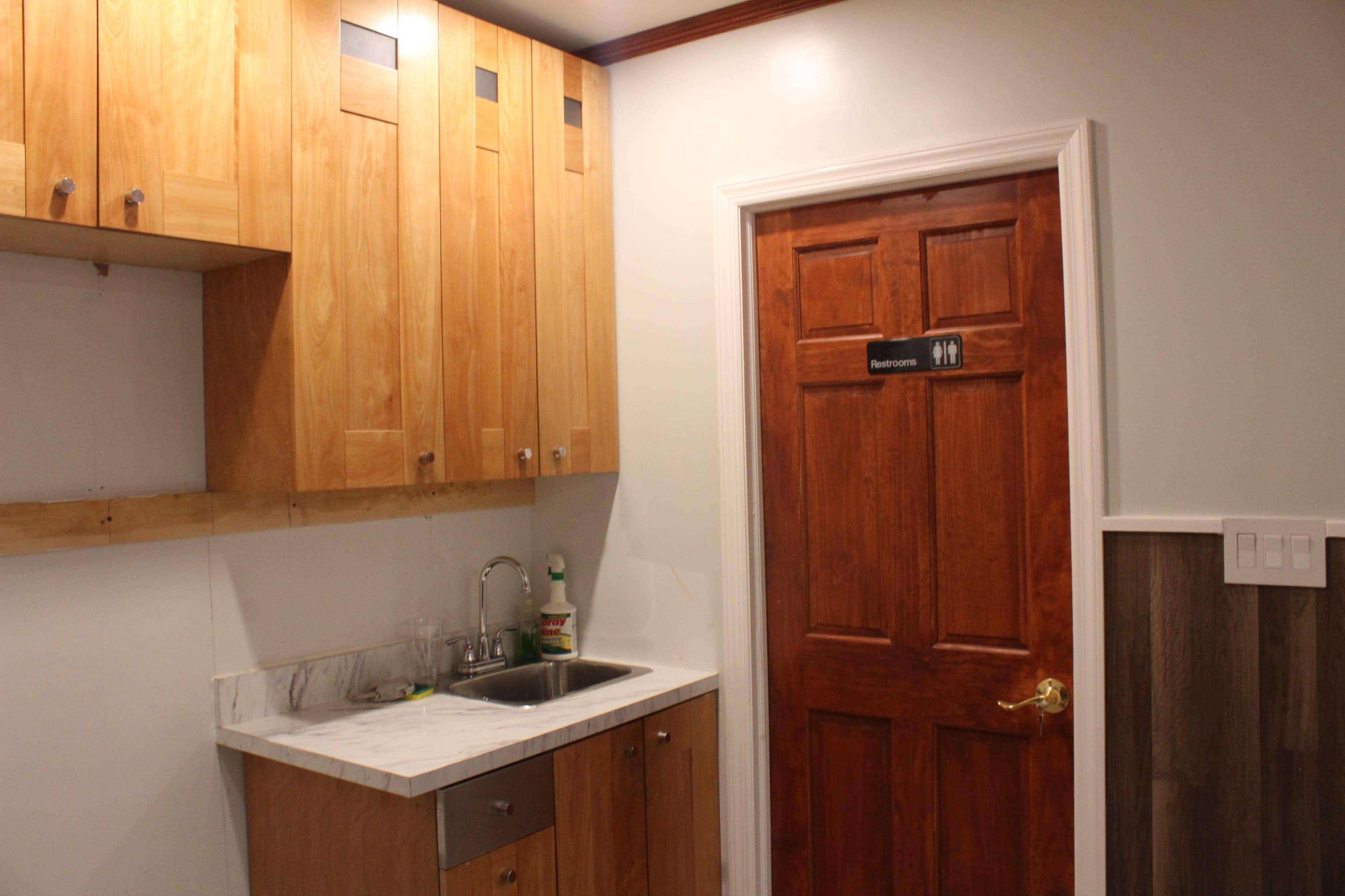 The image shows a small kitchenette area with wooden cabinetry, a stainless steel sink, and a door labeled "Restroom."