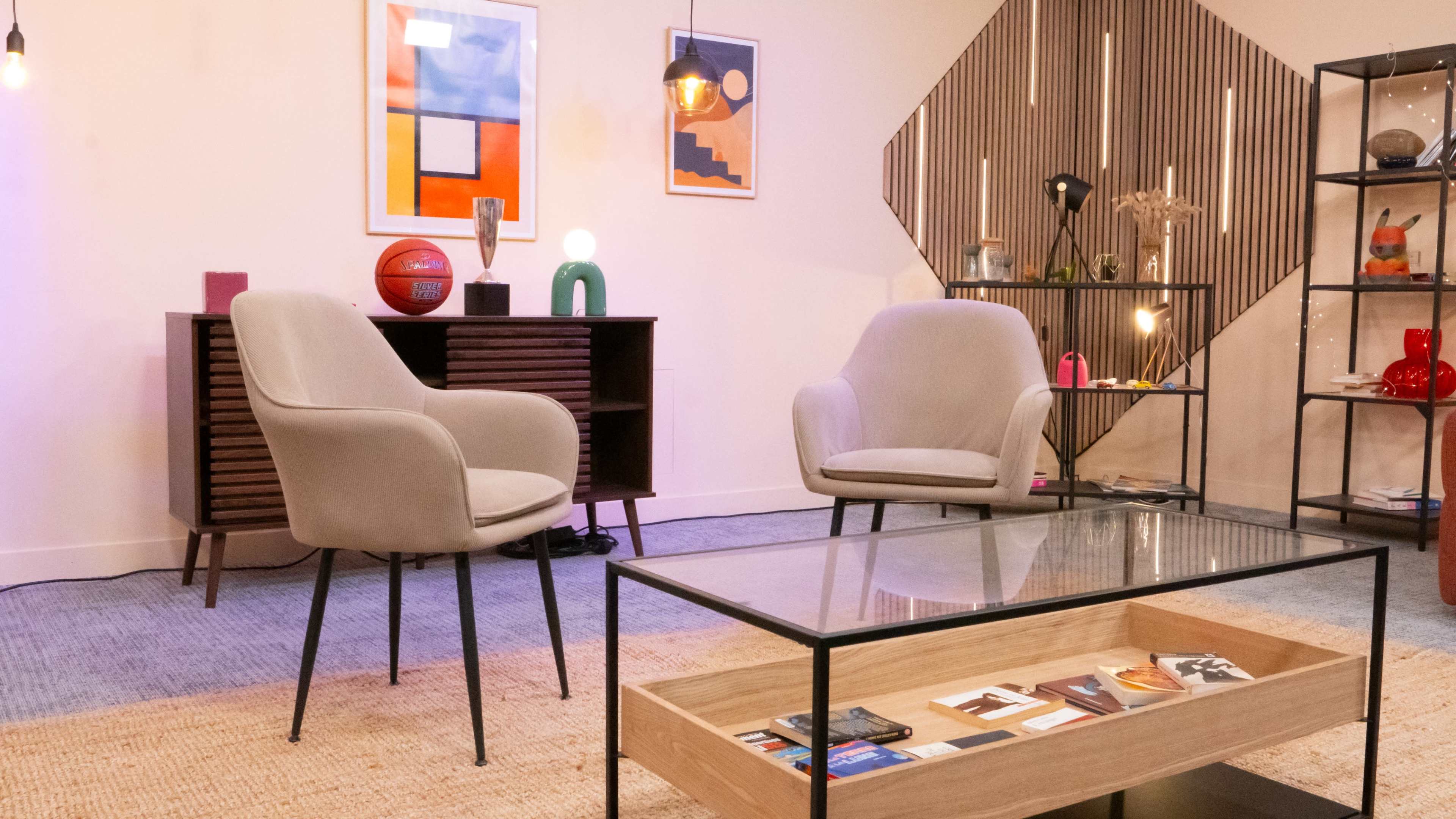 The image shows a modern interior with two chairs facing a glass coffee table, colorful artwork on the walls, and decorative items displayed on shelves.