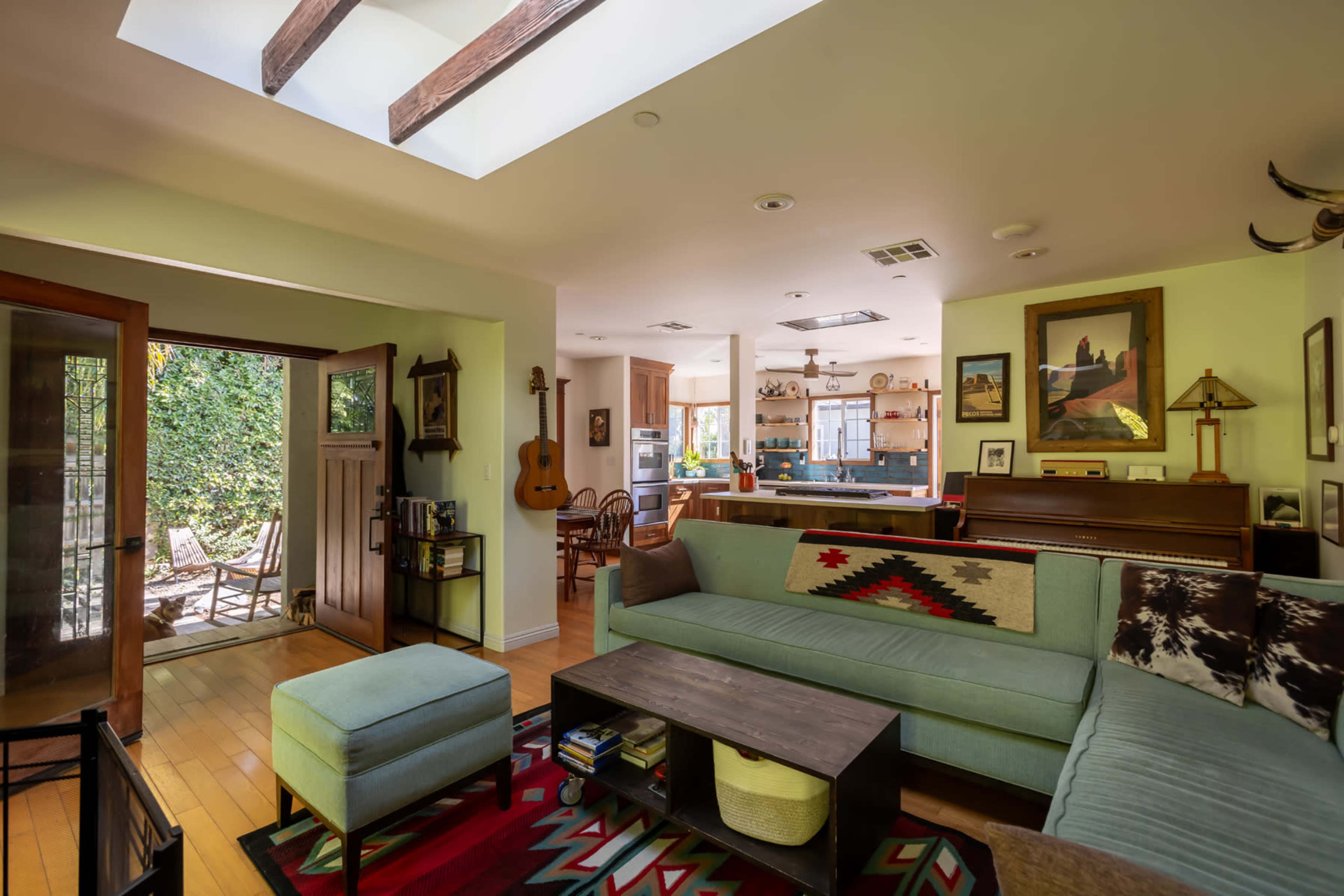 The image shows a cozy living room with a sectional sofa, a wooden coffee table, and an open entryway leading to a patio area, with wooden beams and a skylight above.
