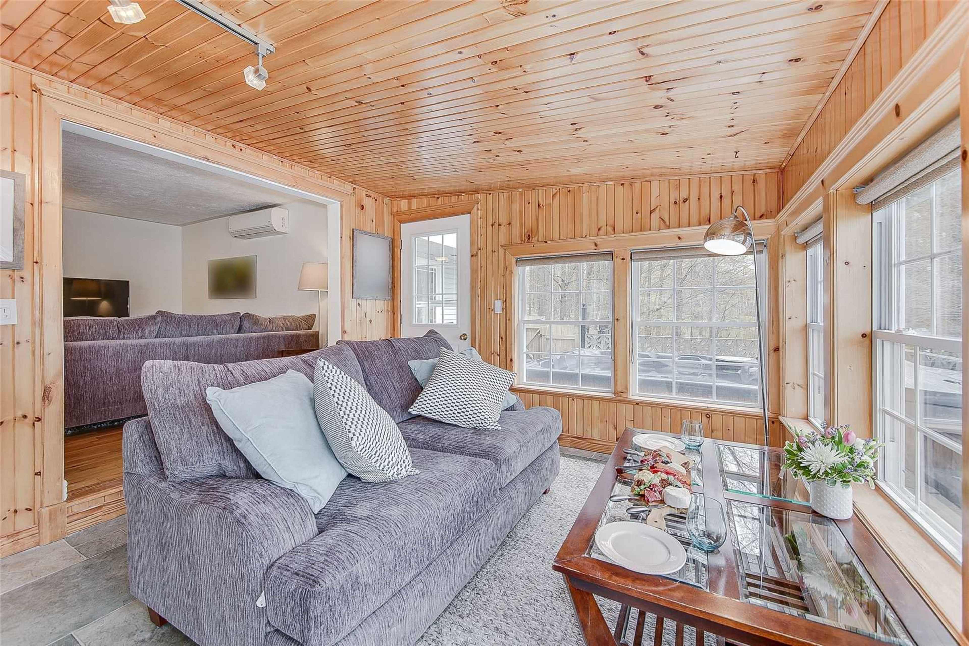 A cozy sitting area features a gray sectional sofa, a glass coffee table set with dishes, and wooden walls, leading into another room with a large couch visible in the background.