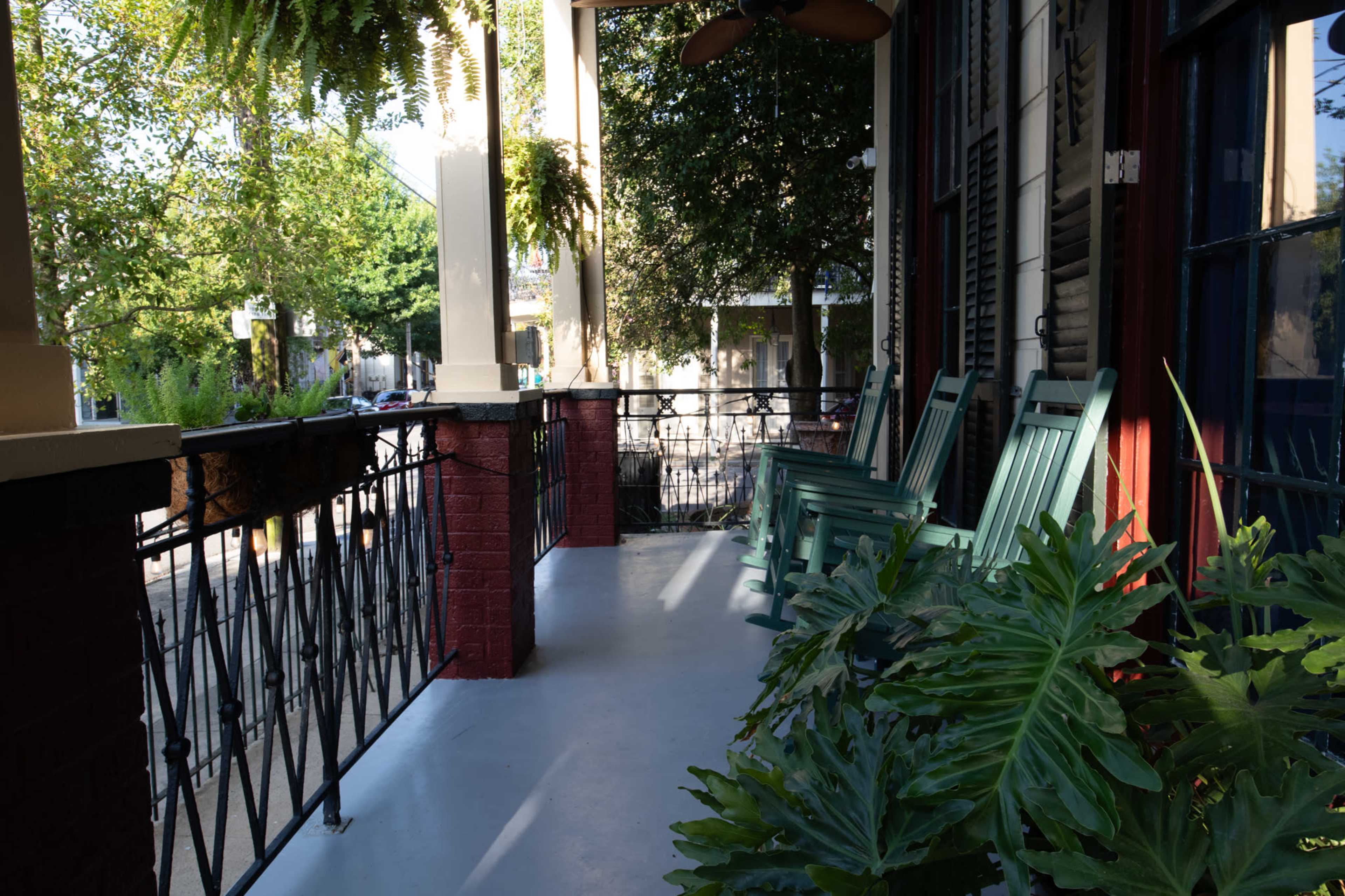 A porch features green rocking chairs alongside a railing, with potted plants and trees visible in the background.