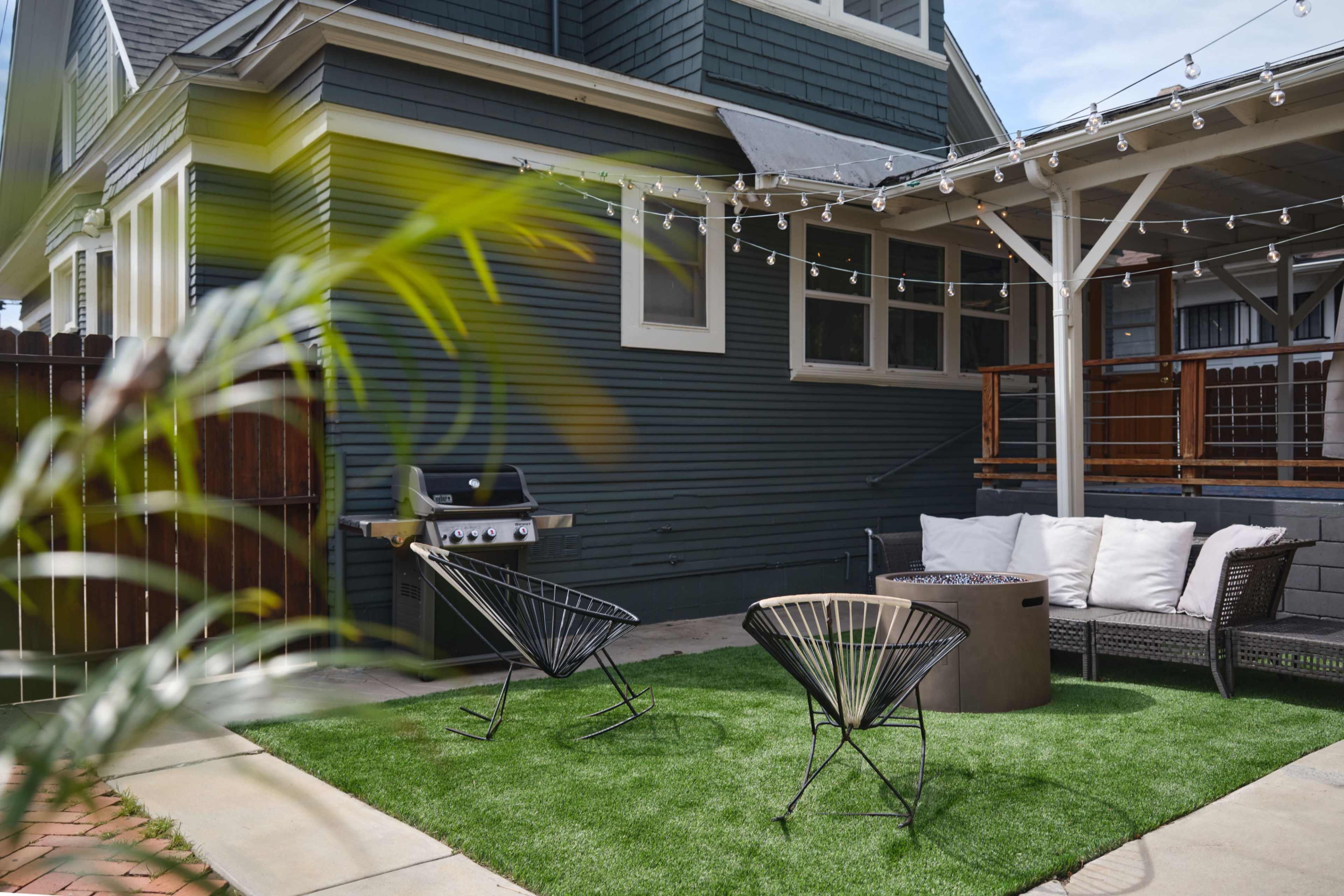 The image depicts a backyard area featuring a barbecue grill, two black patio chairs, a round table, and string lights overhead, all set on a grassy lawn beside a blue house.