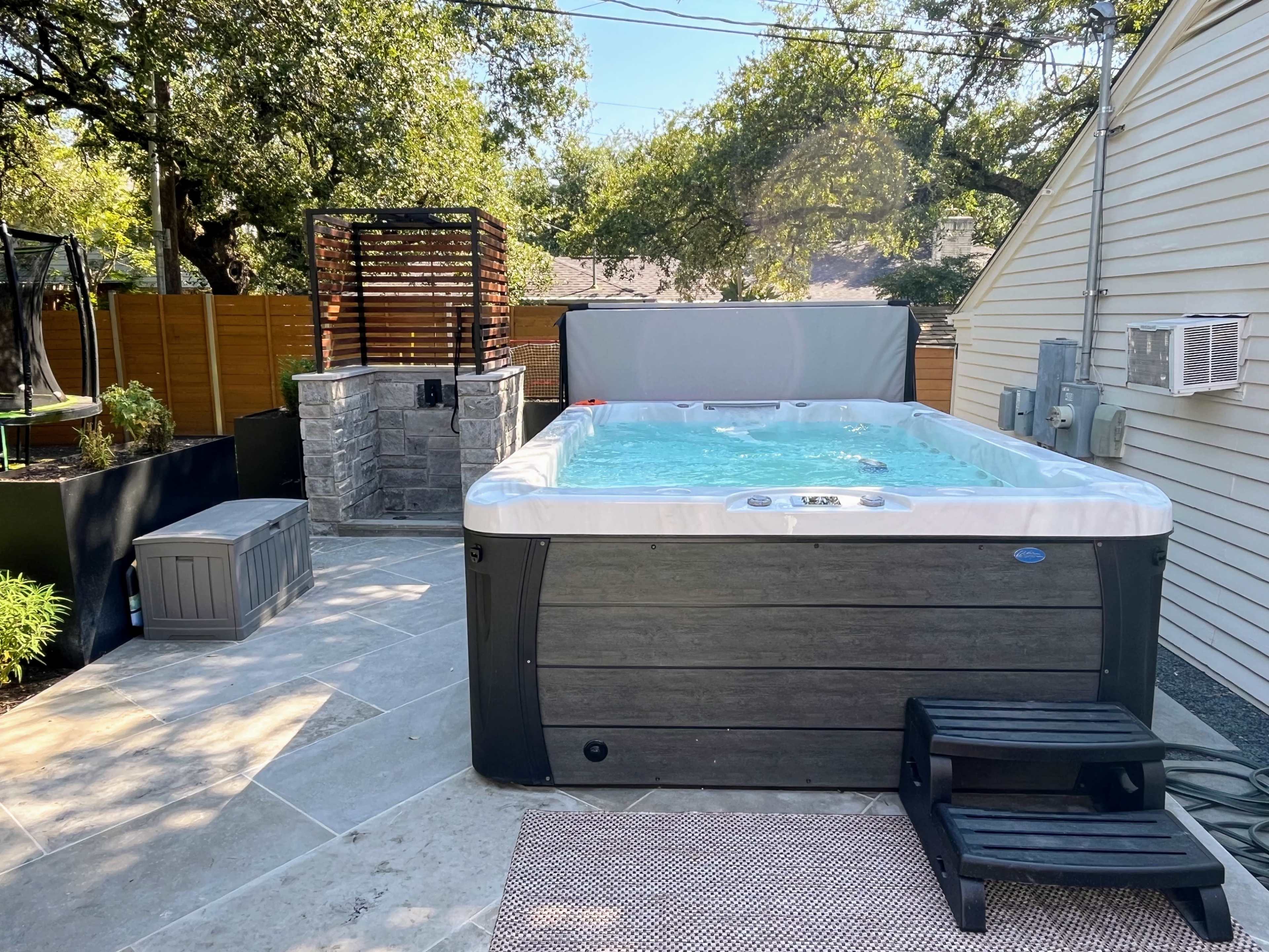 A rectangular hot tub situated on a stone patio, surrounded by greenery and a wooden privacy fence.