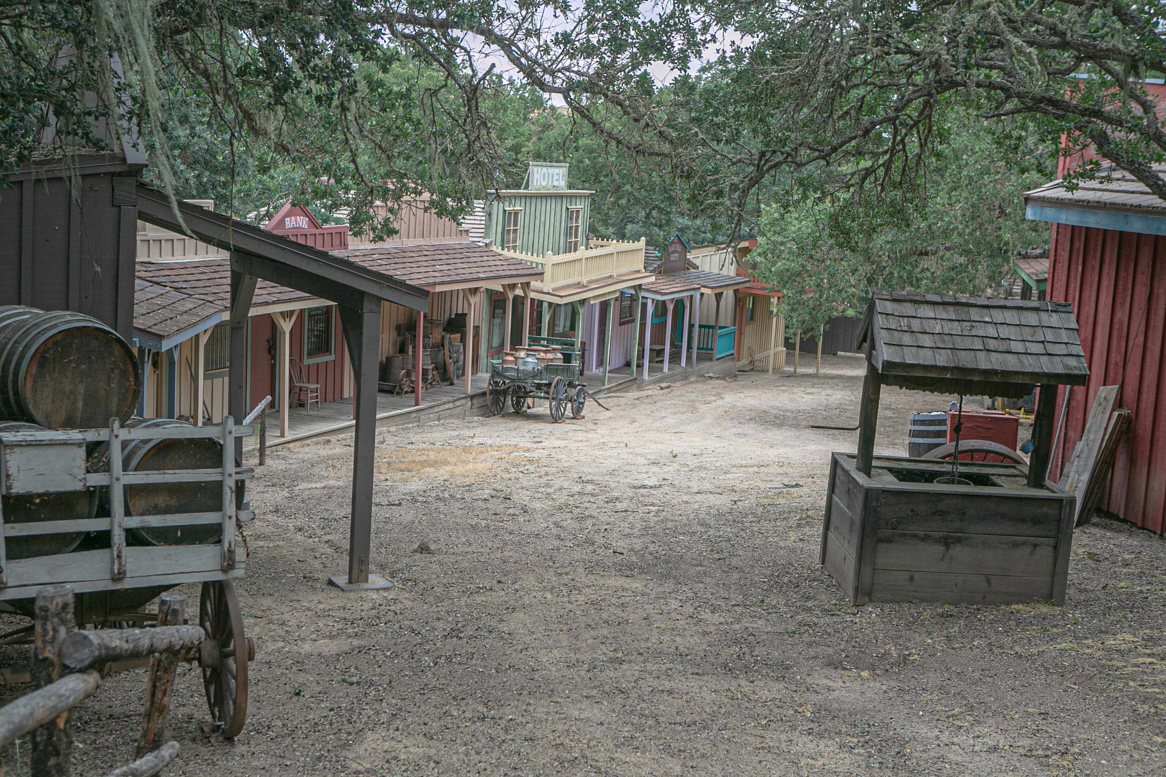 A dirt street lined with old storefronts and a few wooden wagons under a canopy of trees.