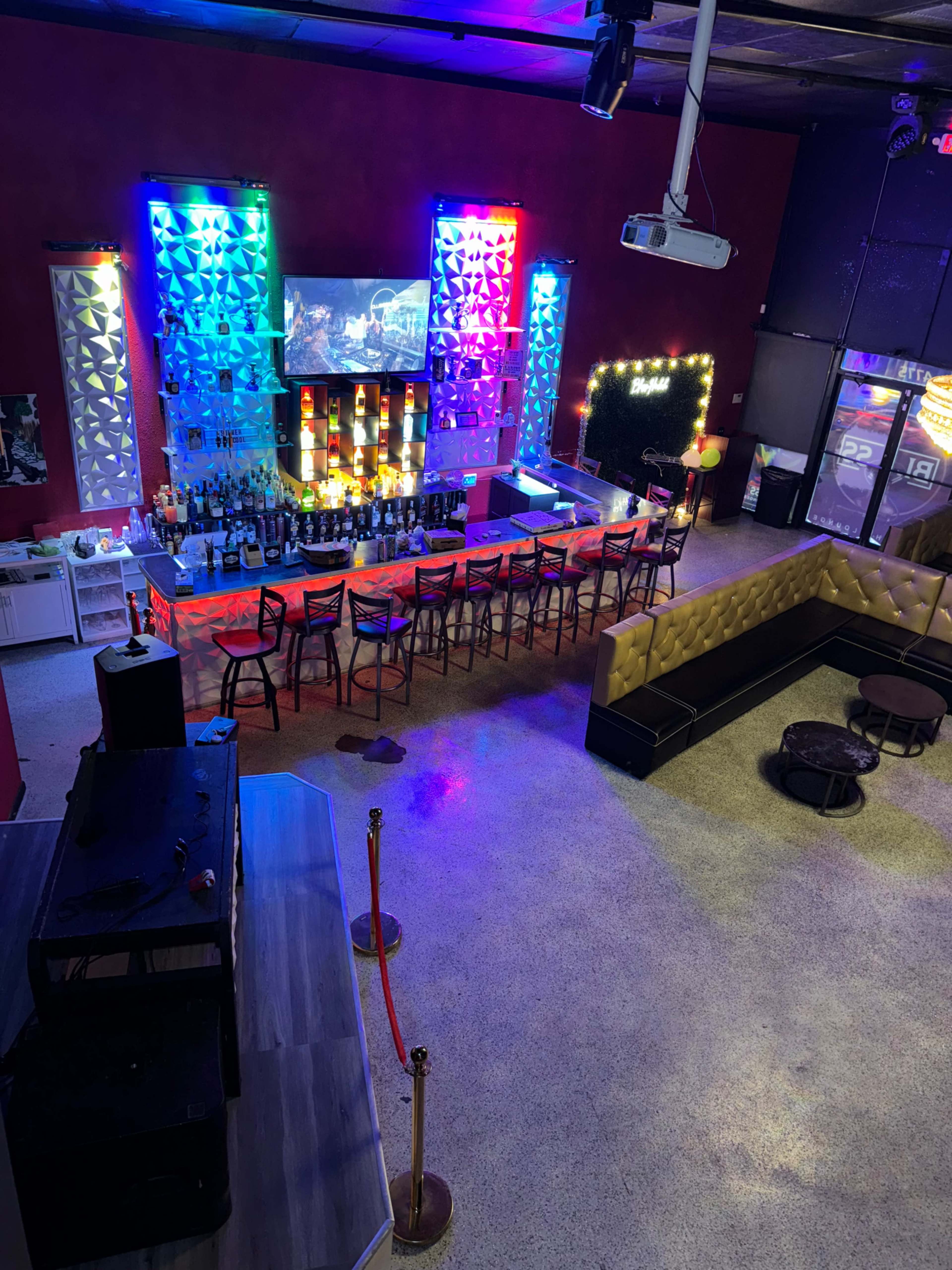 A brightly lit bar area with colorful lights, a linear bar counter lined with various bottles, and a seating section featuring black couches and gold accents.