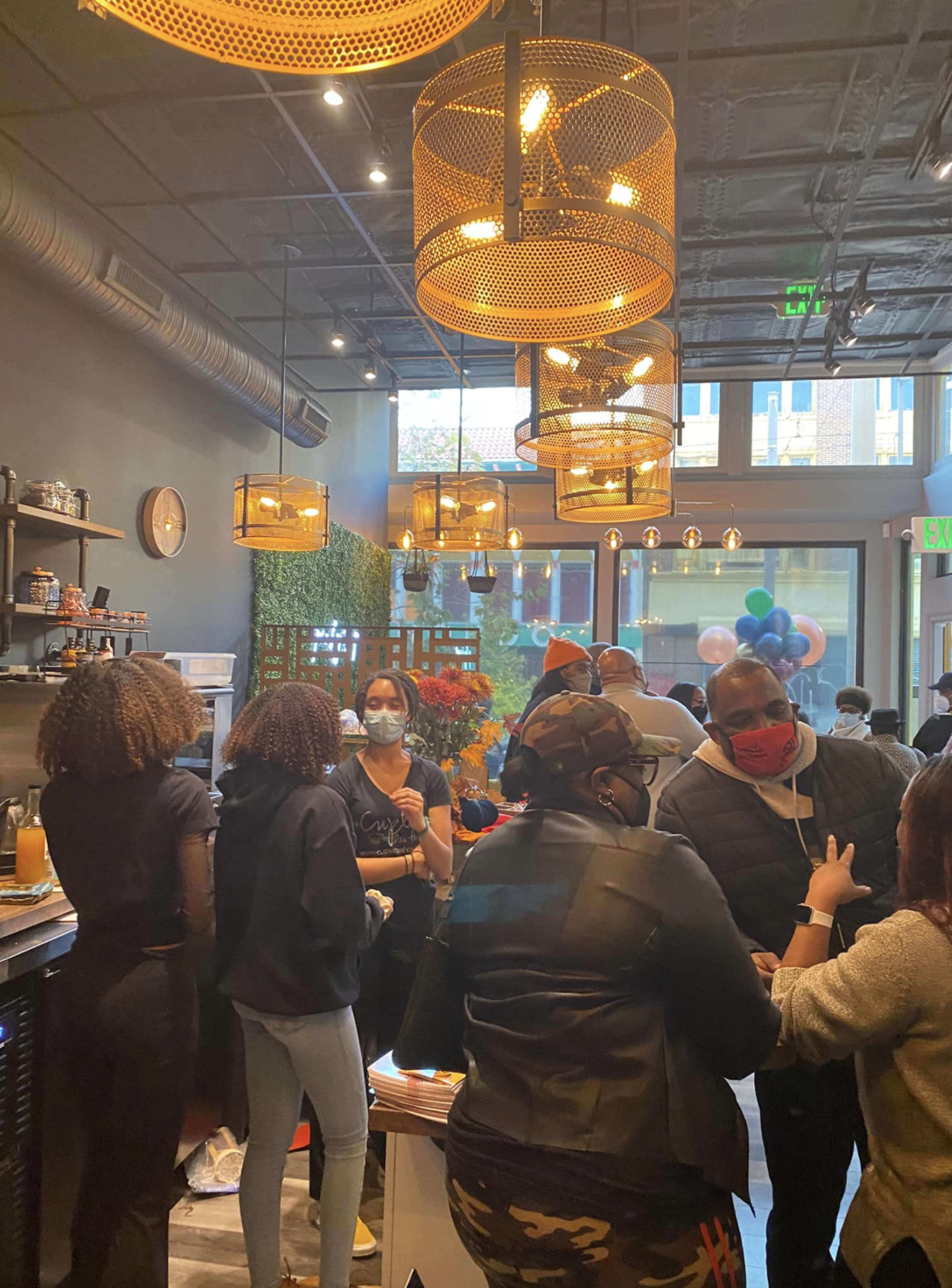 A bustling café interior features people socializing, colorful decor, and hanging pendant lights.