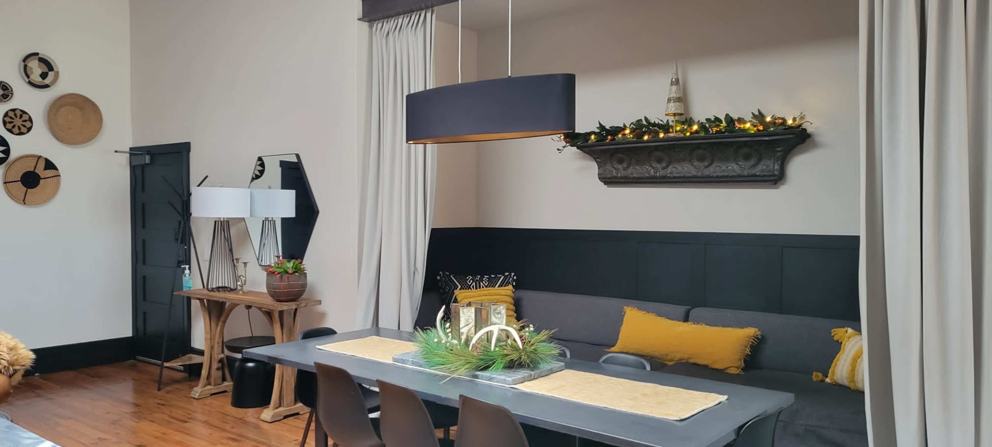 The image shows a modern living space featuring a gray sofa, a long dining table with decorative centerpieces, and a wall adorned with woven wall art.
