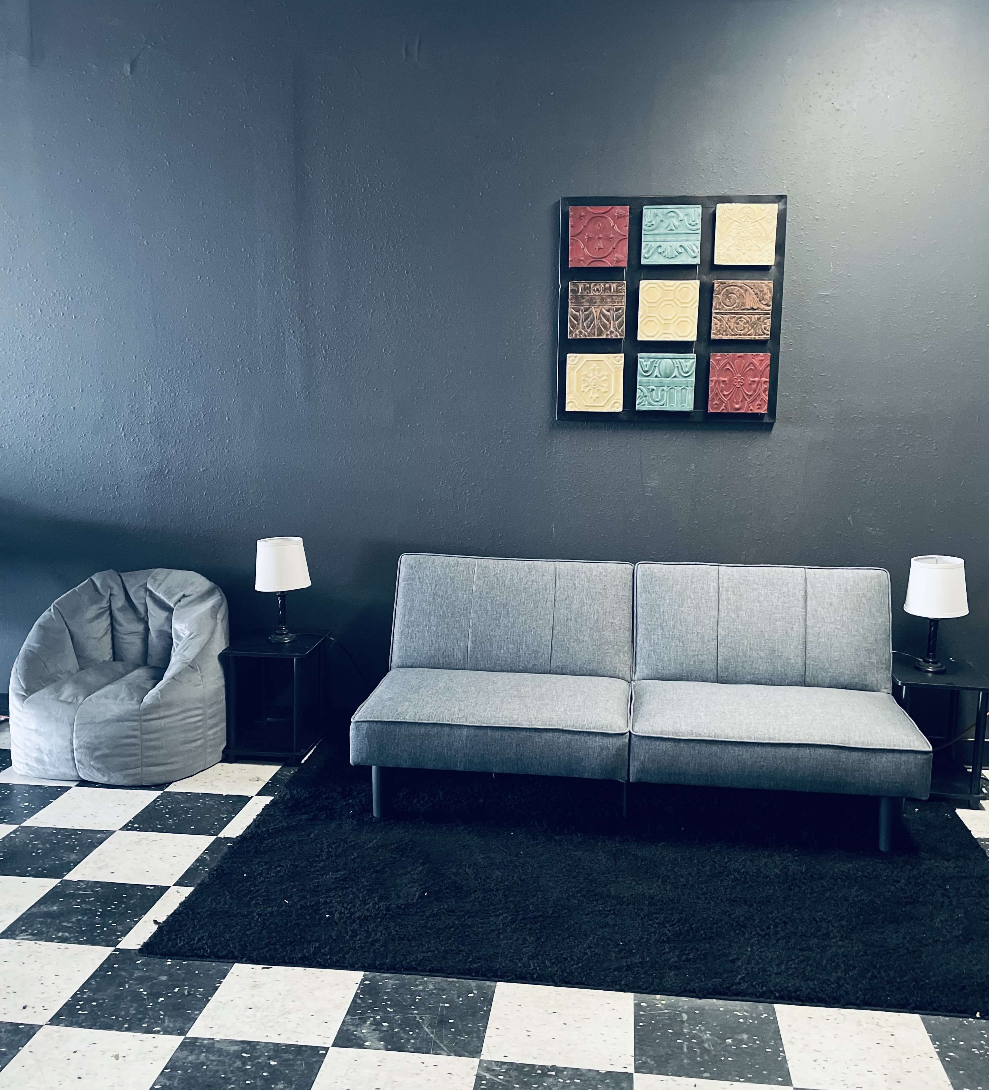 A minimalist seating area with a gray sofa, a bean bag chair, two side tables with lamps, and a colorful wall art piece, all set against a dark wall and a black-and-white checkered floor.