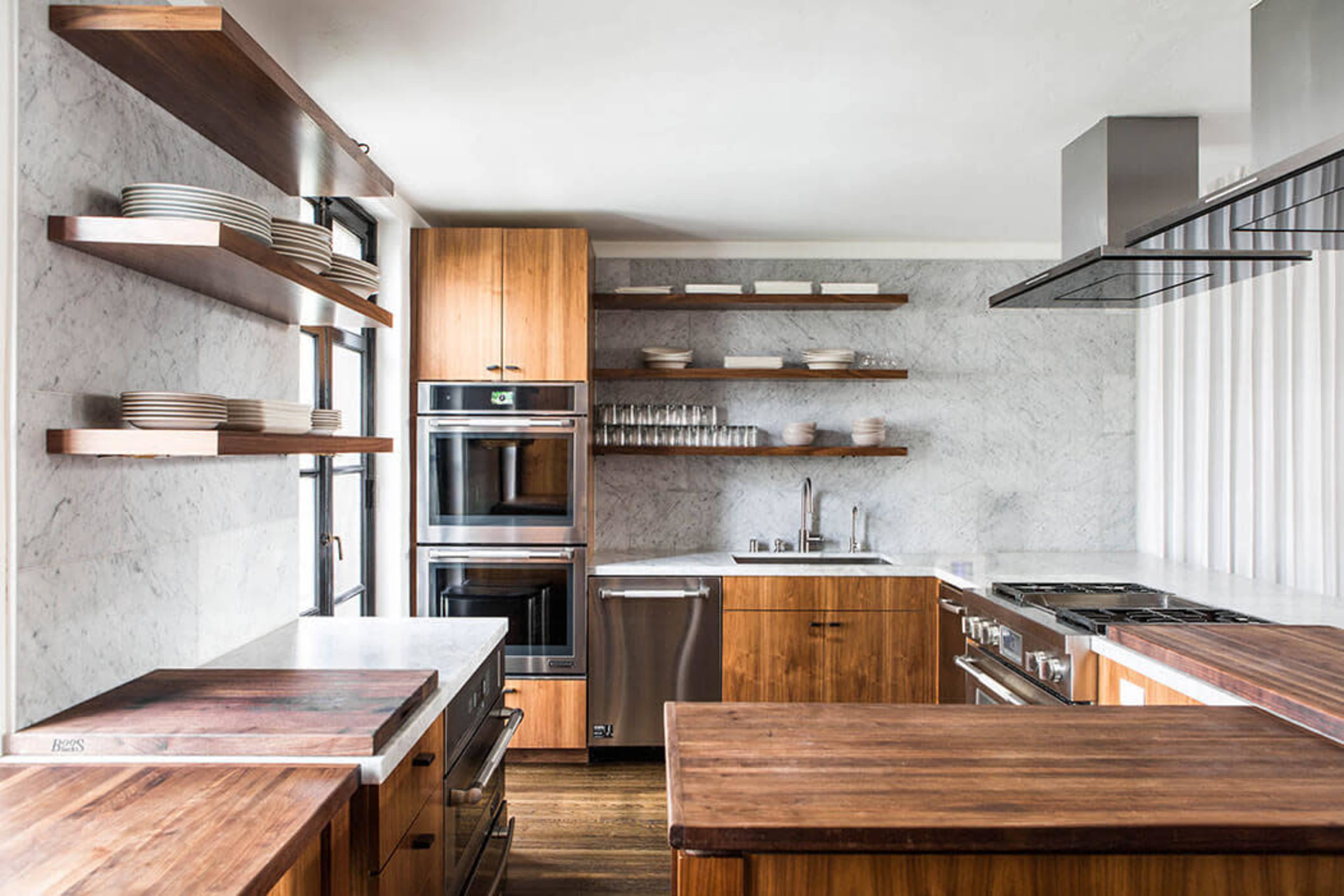 The image shows a modern kitchen featuring wooden cabinetry, open shelving with plates, and stainless steel appliances against a marble backdrop.