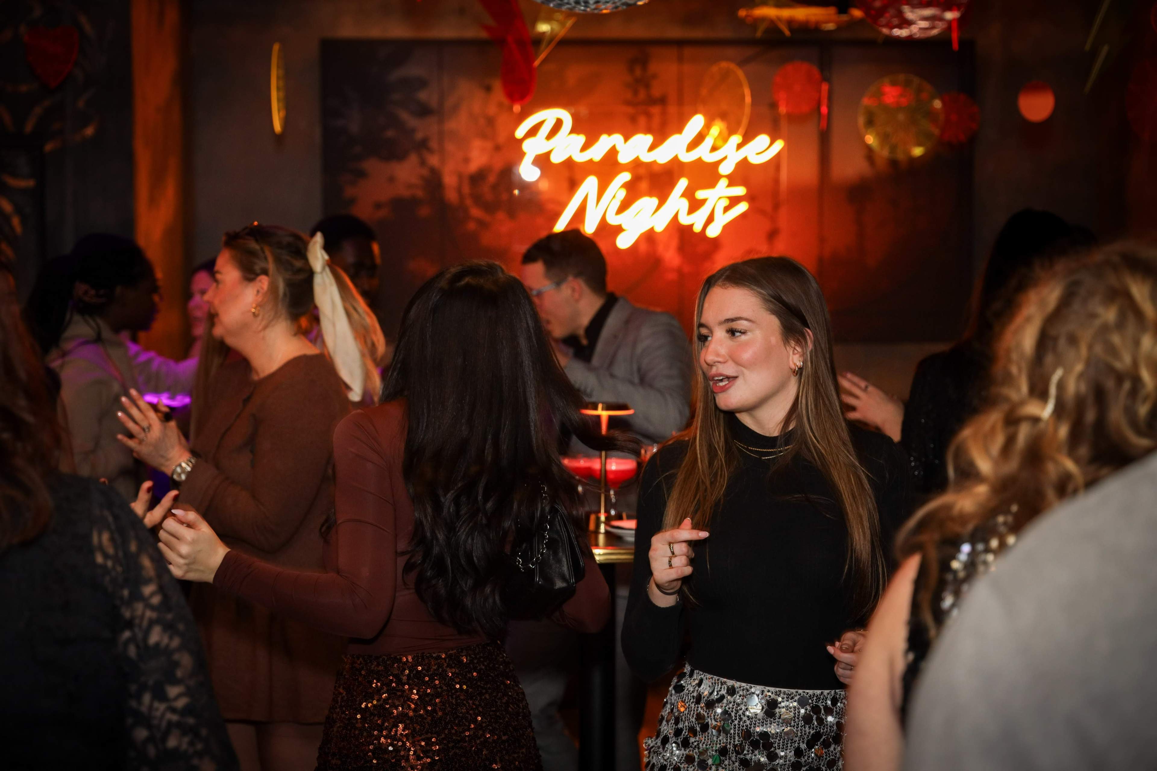 A lively gathering in a dimly lit venue features people dancing and interacting under colorful decorations and a glowing neon sign that reads "Paradise Nights."