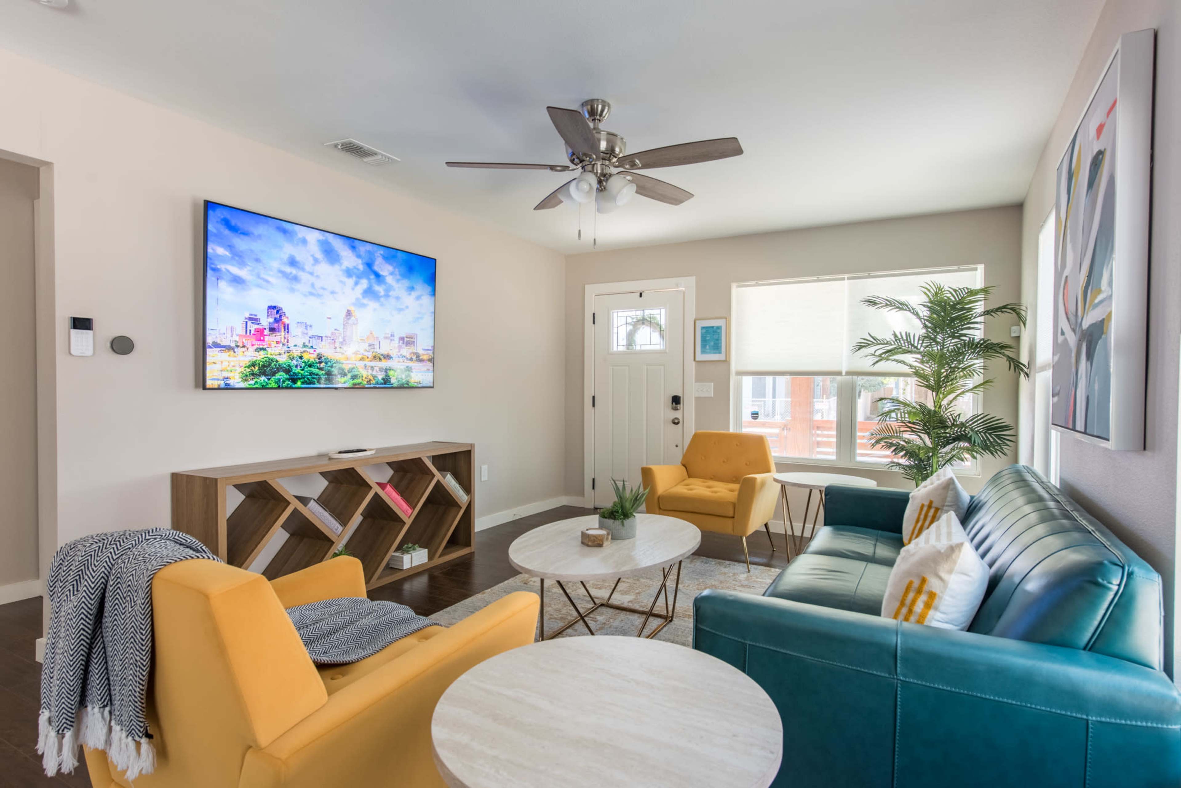 The image shows a modern living room with a large wall-mounted TV, two colorful sofas, a coffee table, and a door leading outside.