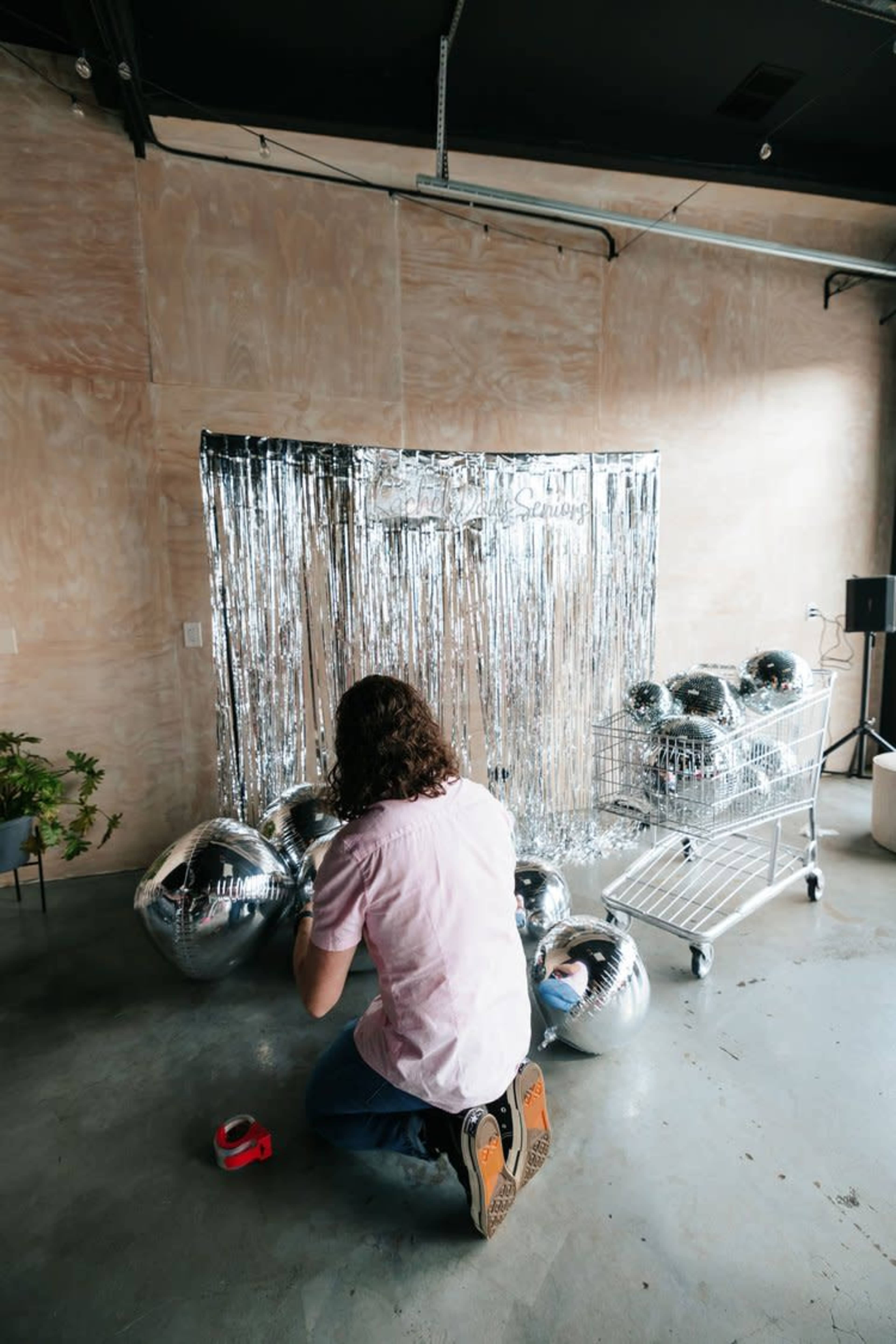 A person kneels on the floor, preparing silver balloons in front of a shiny metallic backdrop in a sparse room.