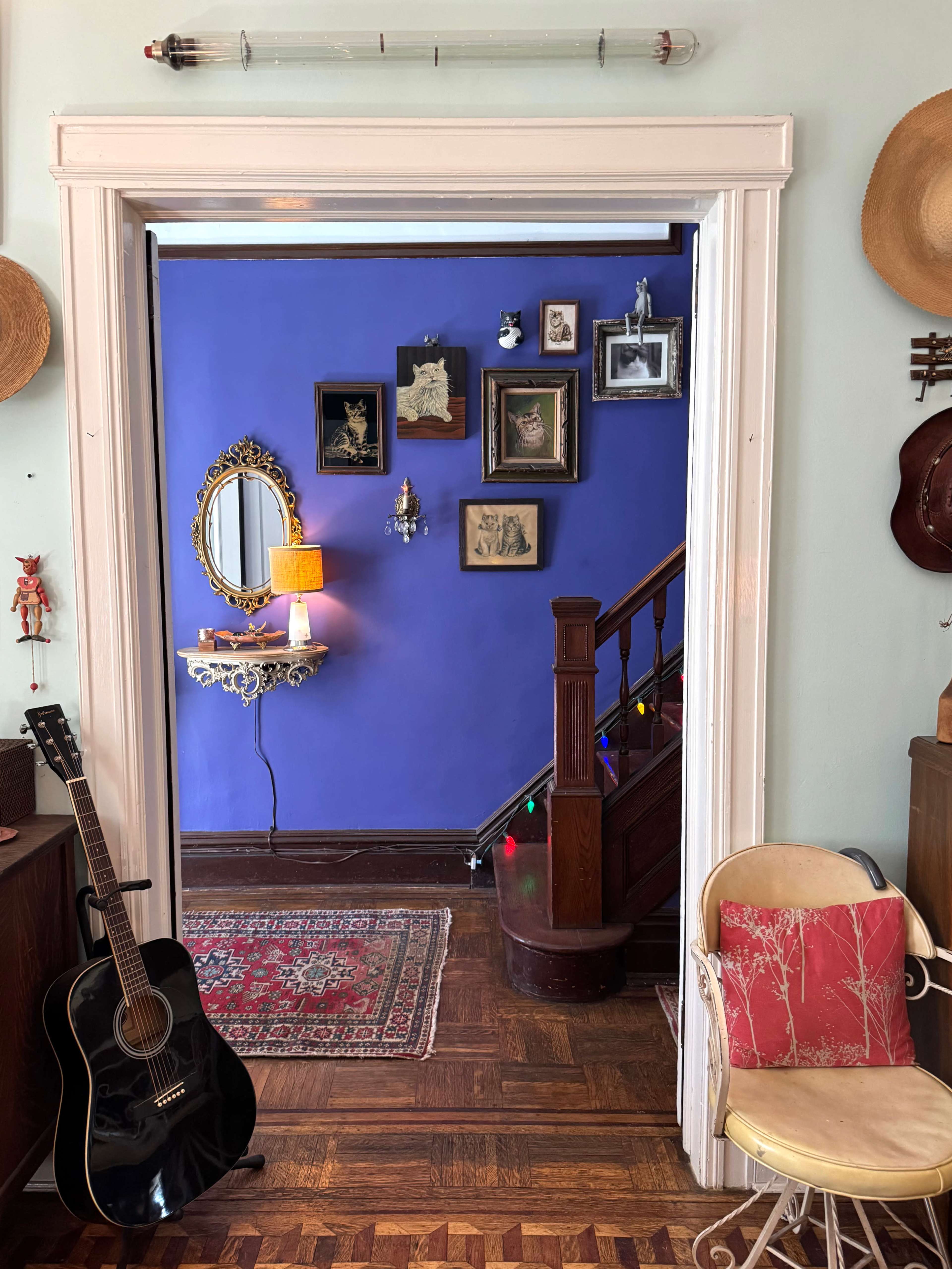 A hallway features a blue wall adorned with framed pictures, a mirror, and a staircase, while a guitar leans against a nearby wall and a chair with a cushion sits to the side.