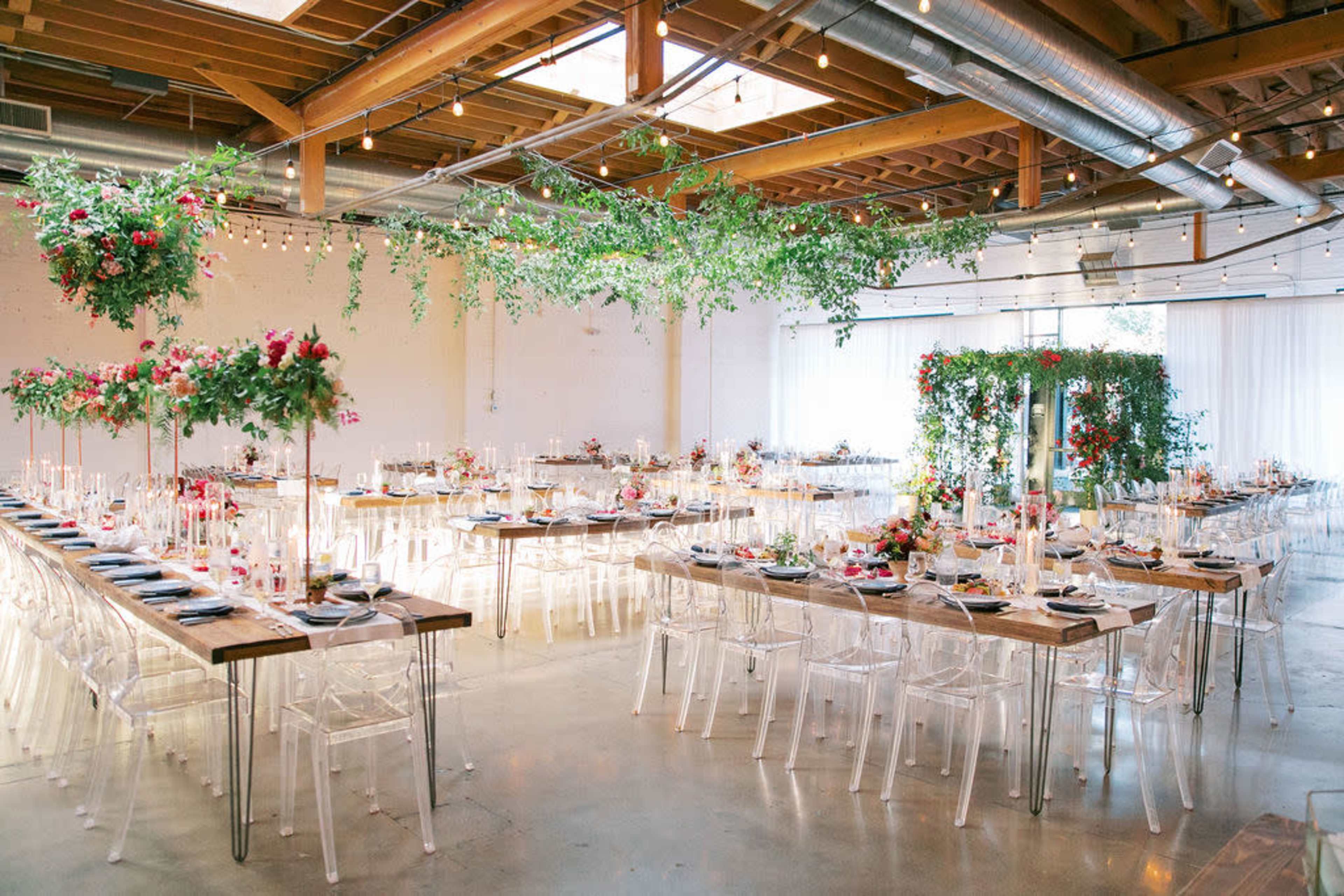 A well-decorated event space with long dining tables set for a gathering, featuring floral arrangements and string lights overhead.