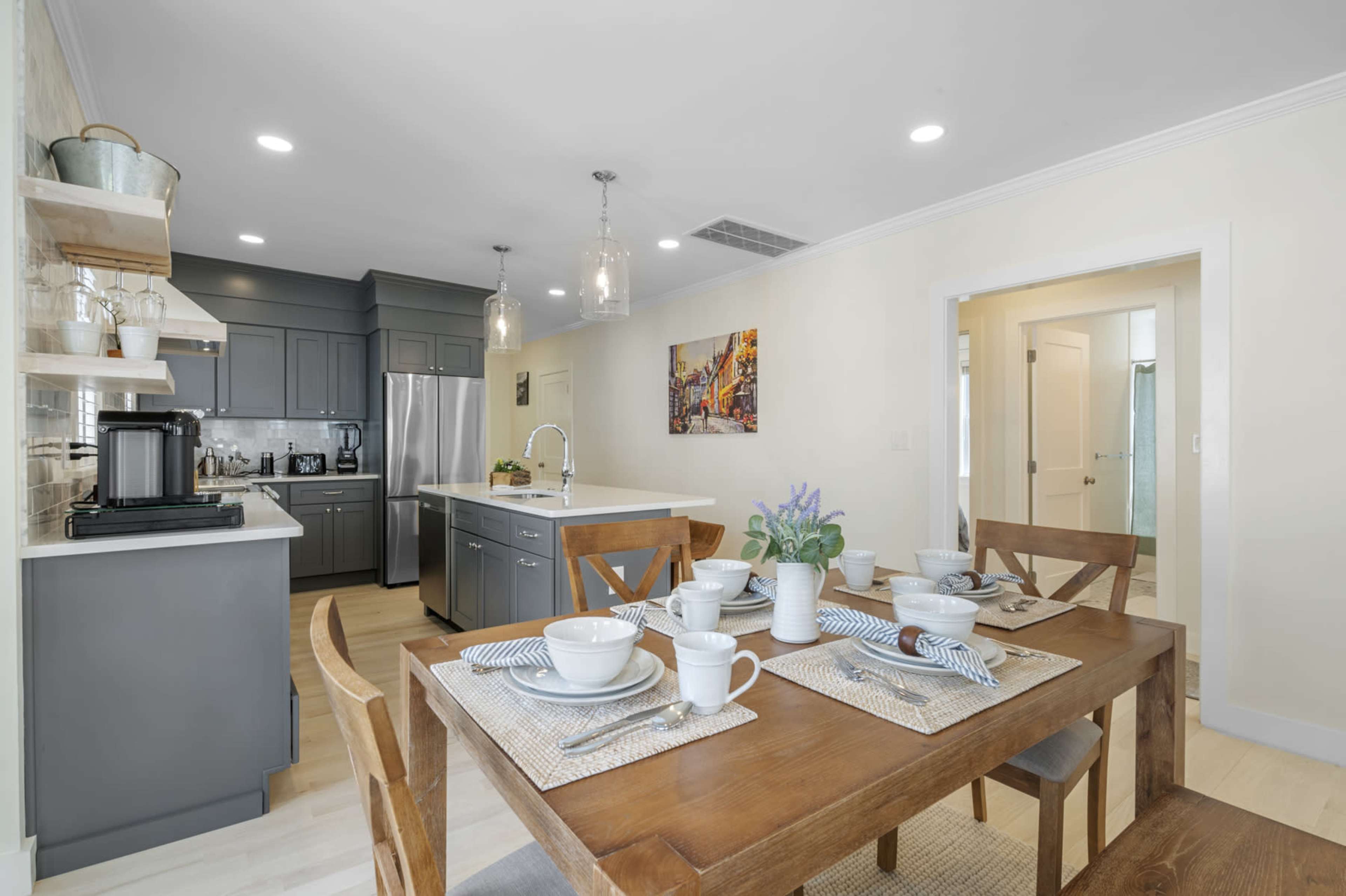 A dining area with a wooden table set for four, adjacent to a modern kitchen featuring gray cabinets and stainless steel appliances.