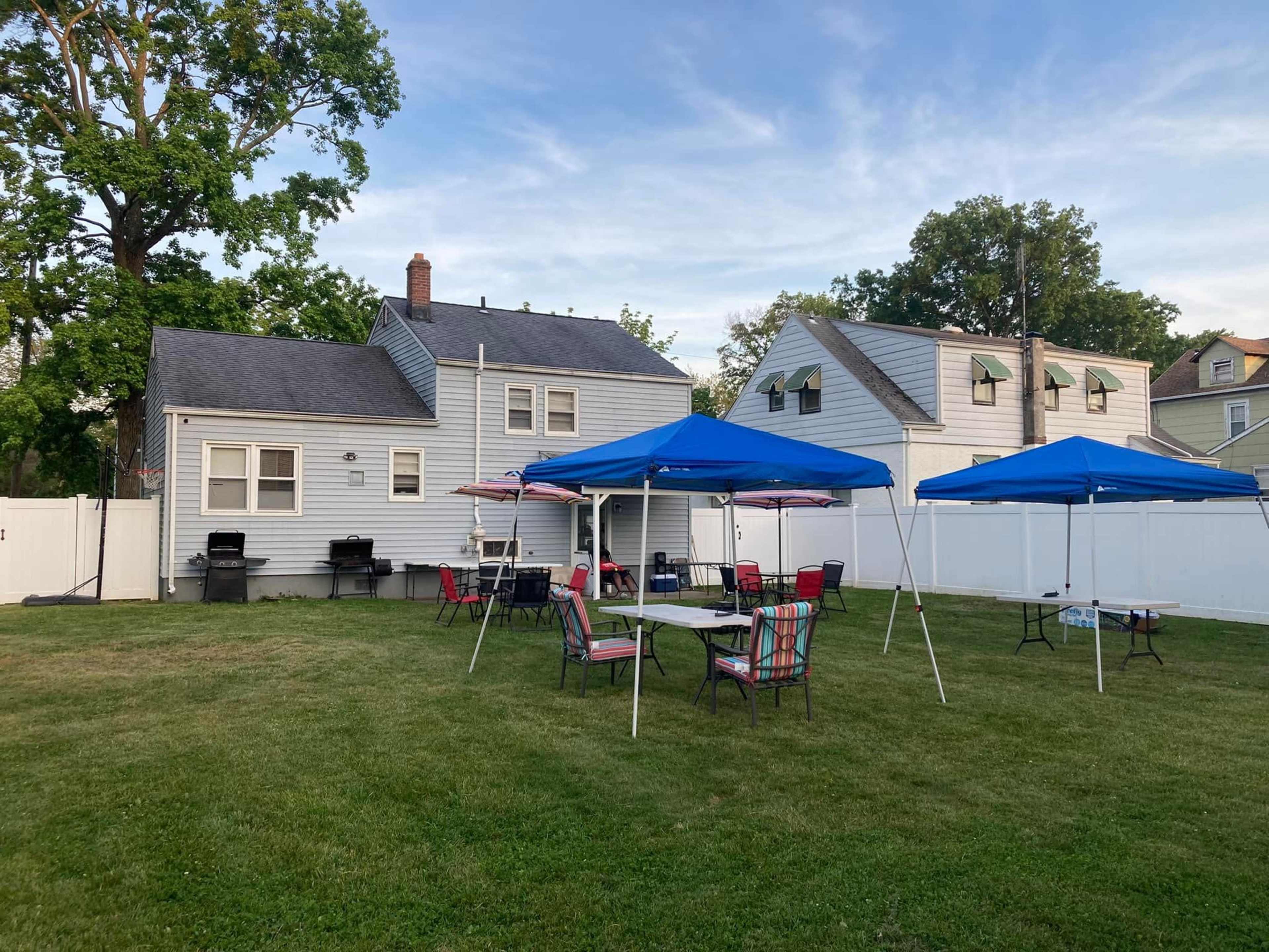 A backyard features several blue canopies set up over tables and chairs, with a house and a white fence in the background.