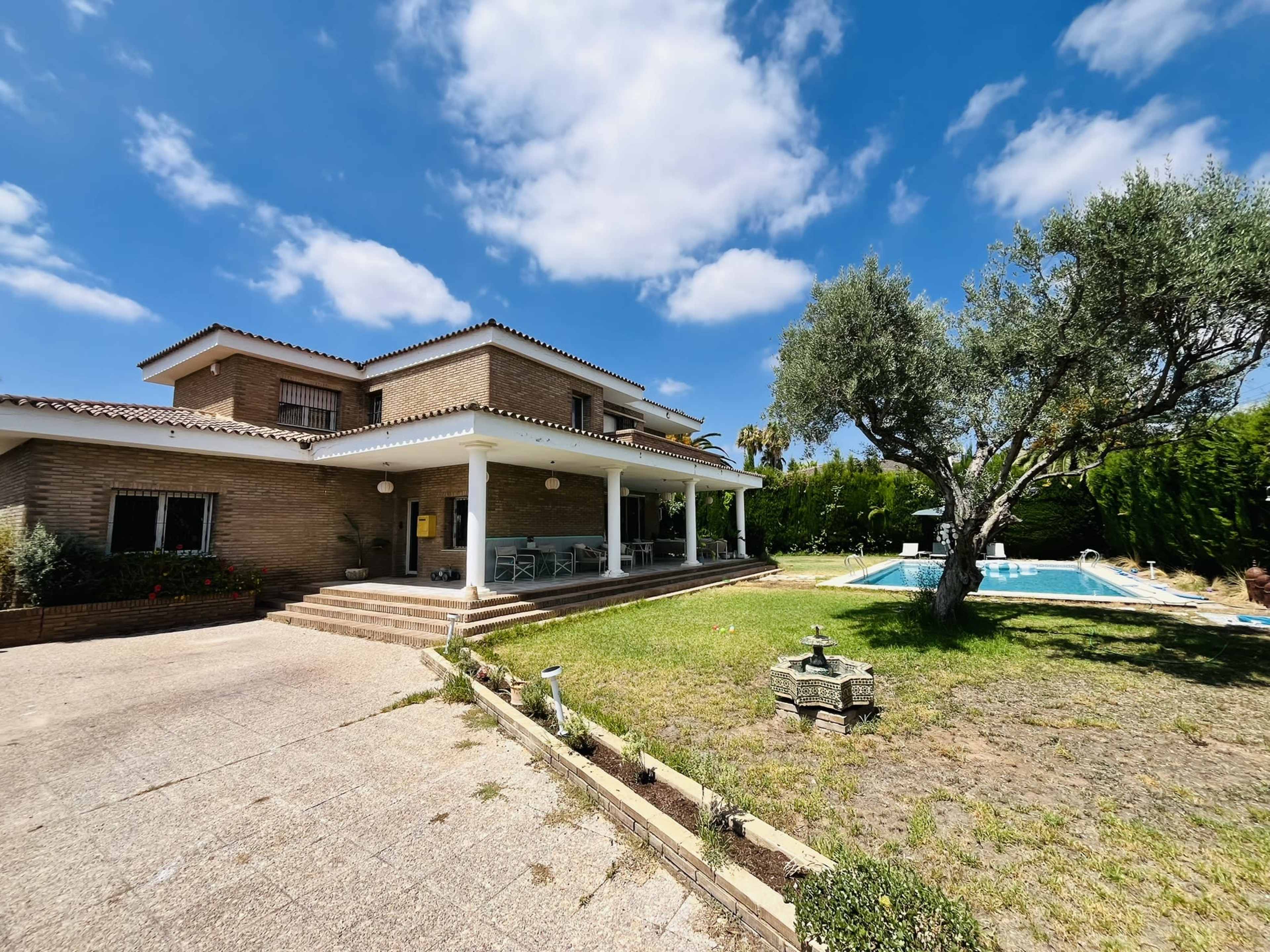 The image shows a large two-story house with a patio area, surrounded by a grassy yard and a swimming pool, under a partly cloudy sky.
