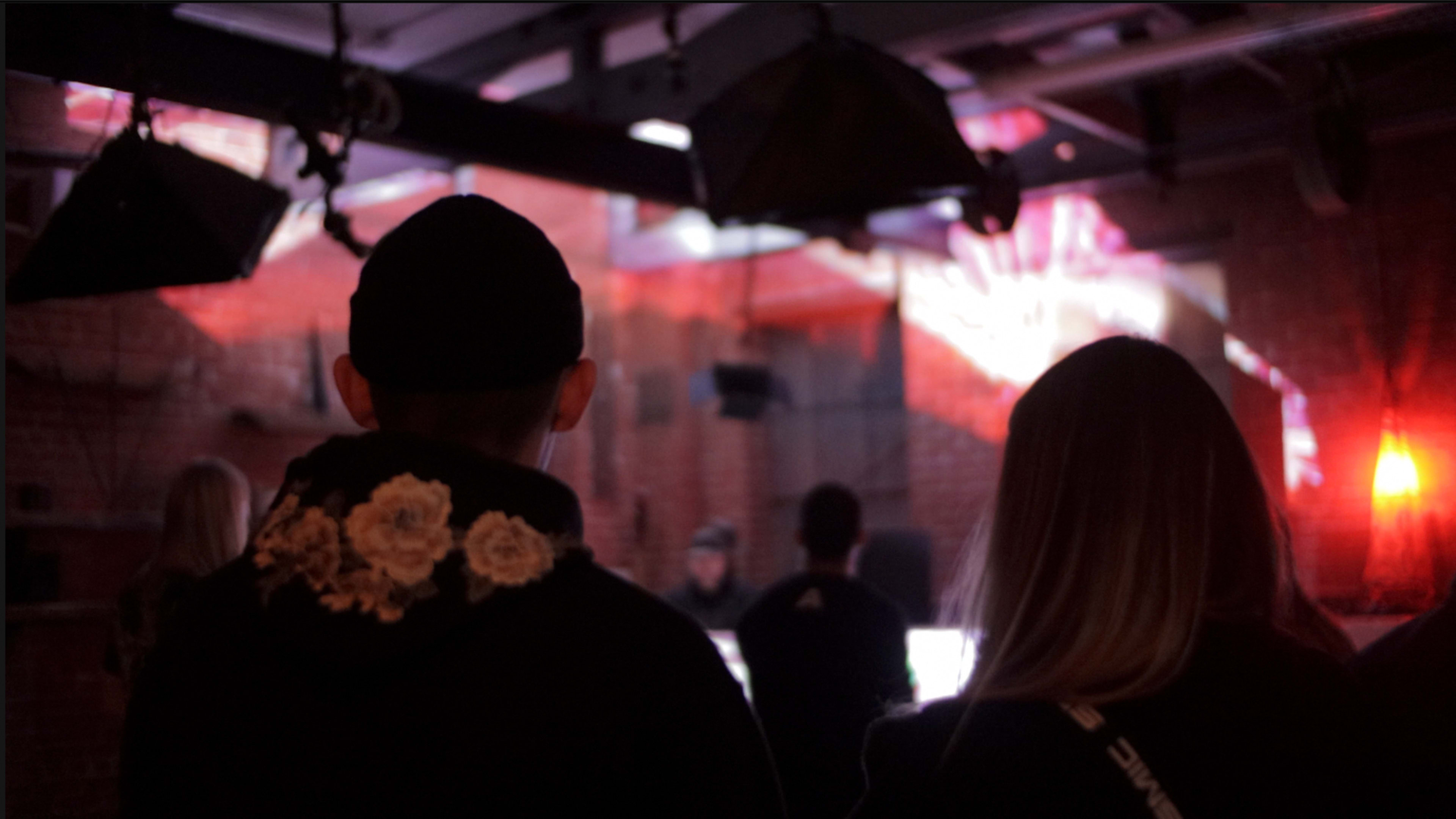 Two people watch a performer in a dimly lit venue with colorful lighting effects.