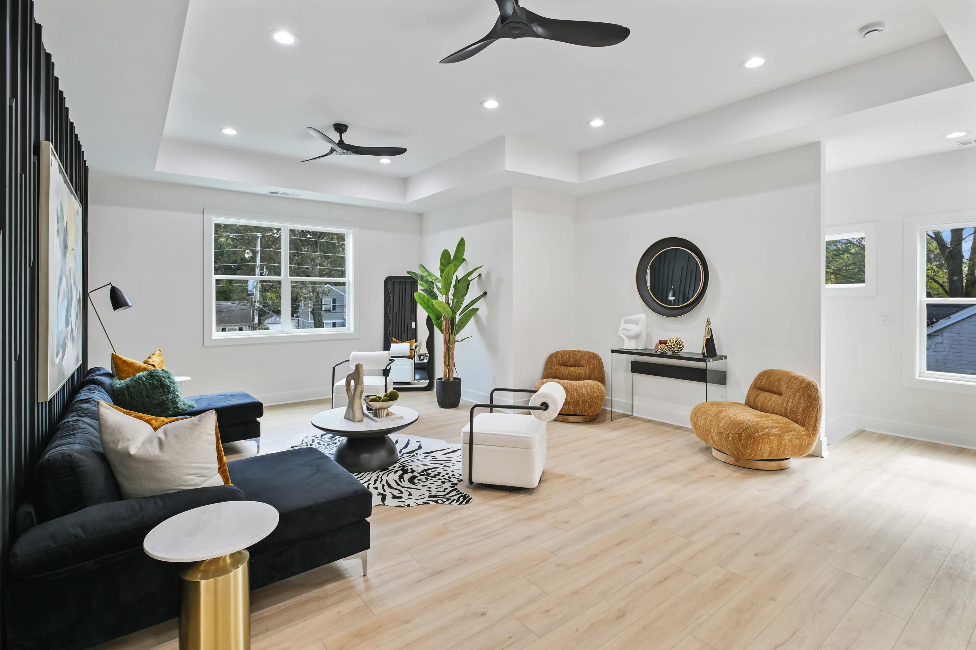The image shows a modern, well-lit living room featuring a black sofa, two brown accent chairs, a circular coffee table, and a decorative black wall.