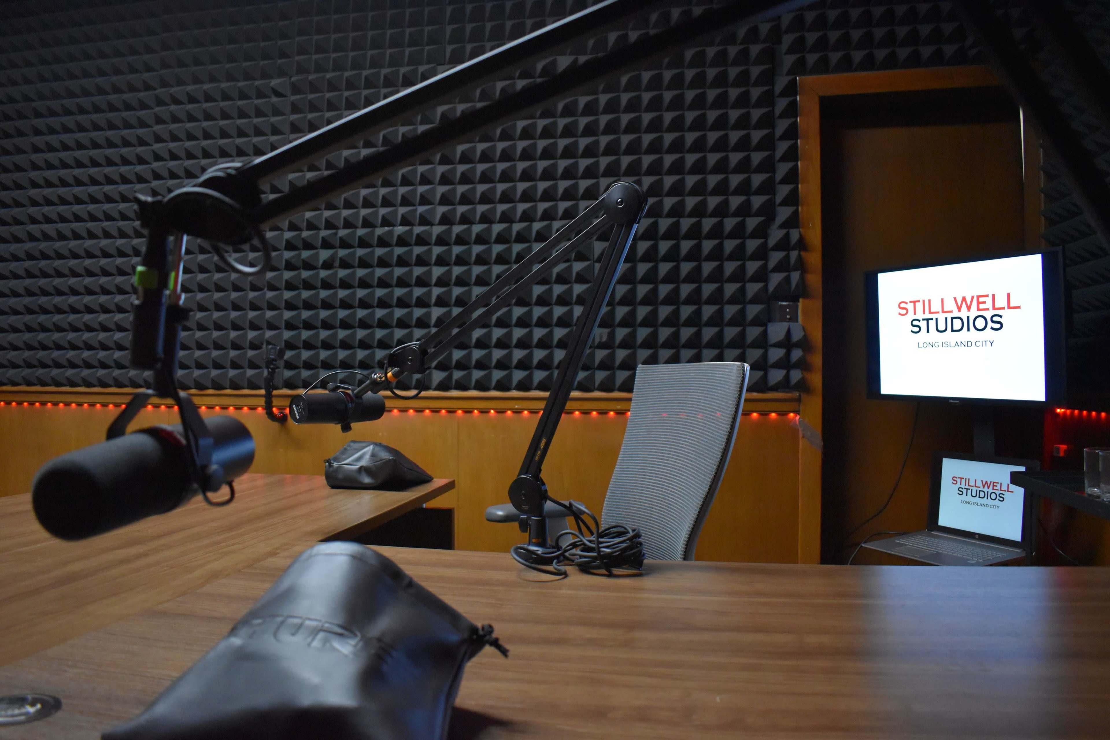 A recording studio with microphones on adjustable arms, a wooden table, and a screen displaying "STILLWELL STUDIOS" in a dimly lit room with soundproof walls.