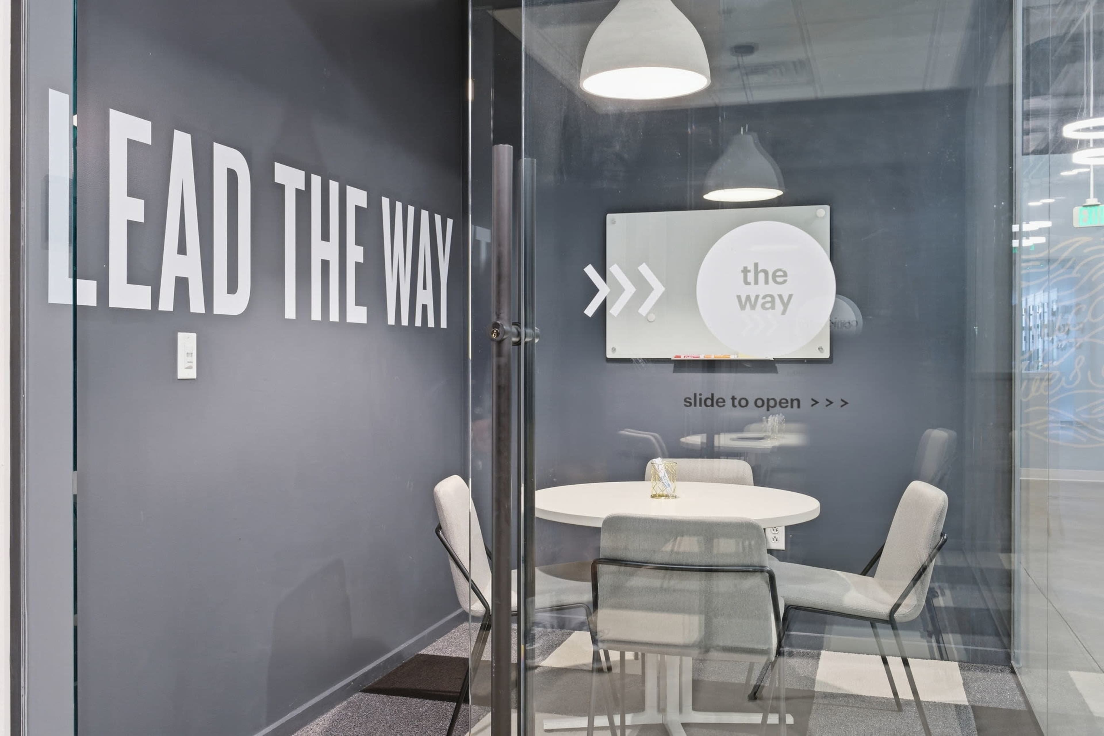 The image shows a modern meeting room with a round table and four chairs, featuring glass walls and a wall with large text that reads "LEAD THE WAY."