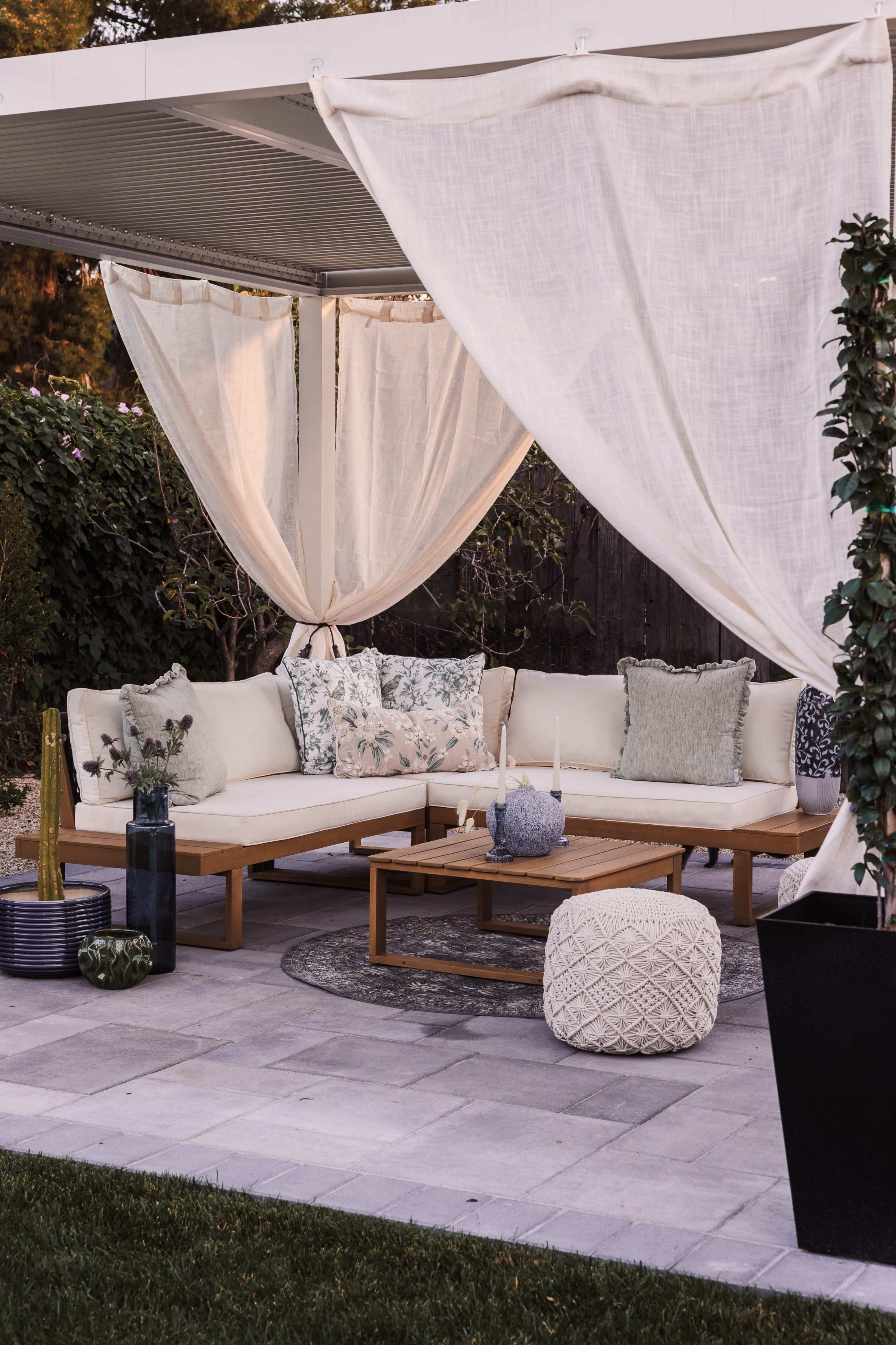 A patio area features a cozy seating arrangement with light-colored cushions, surrounded by sheer curtains and greenery.