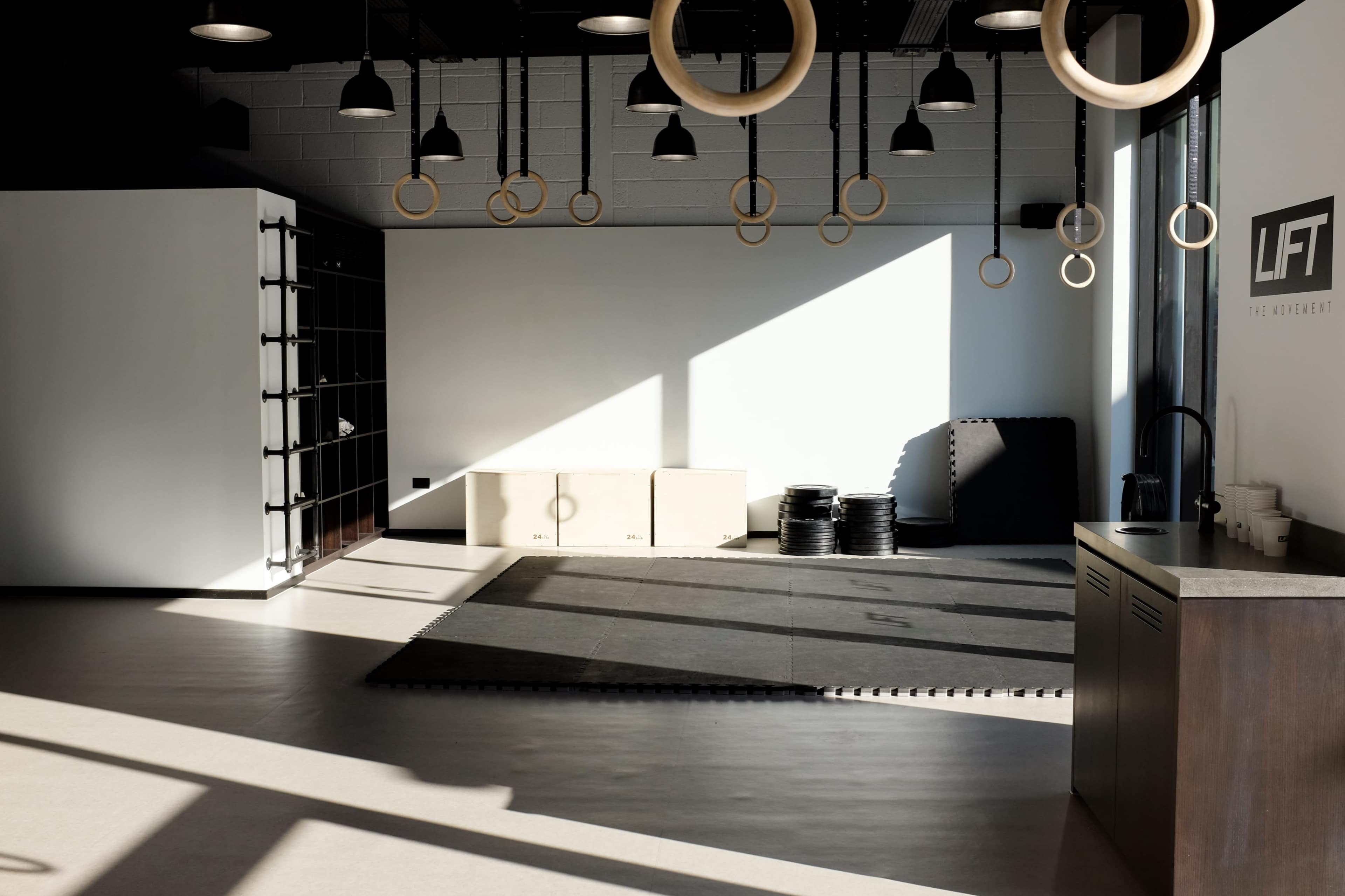 A spacious fitness studio with wooden gymnastic rings hanging from the ceiling, a large window allowing sunlight to cast shadows on the floor, and neatly arranged exercise equipment along the walls.