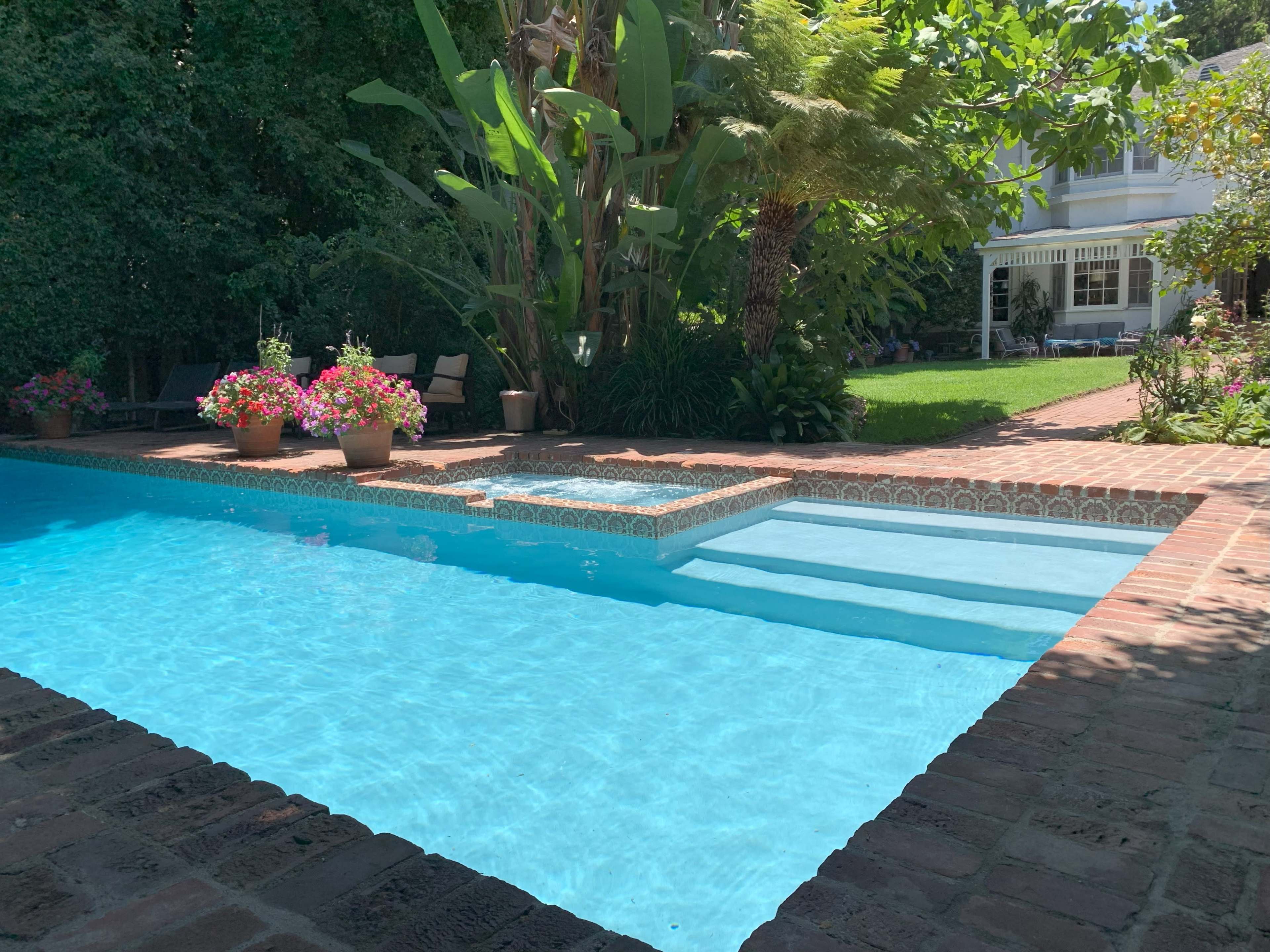 A swimming pool with a hot tub, surrounded by lush greenery and potted flowers, adjacent to a house.