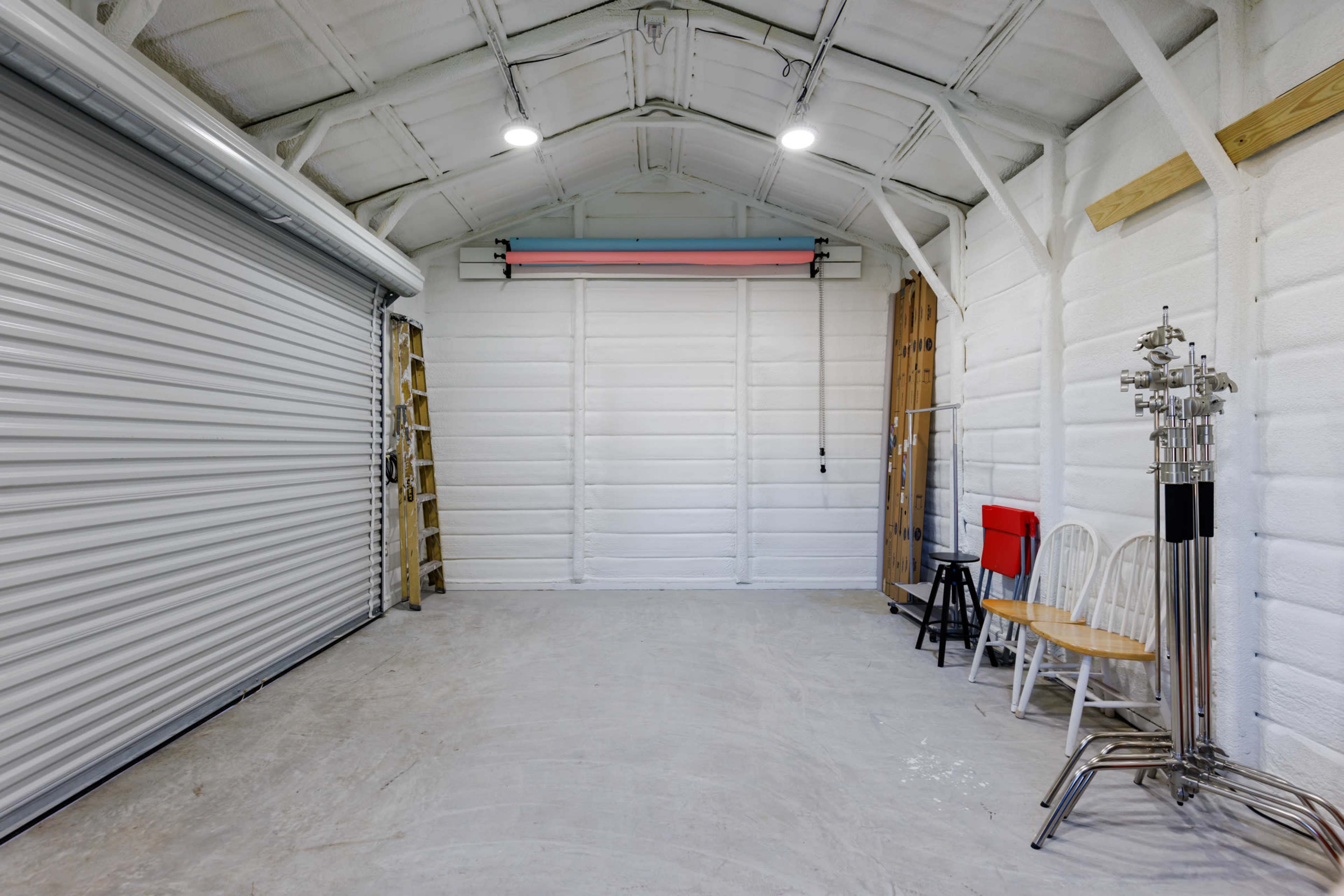 A spacious, empty garage with a roll-up door, several chairs, and ladders against the white-paneled walls.