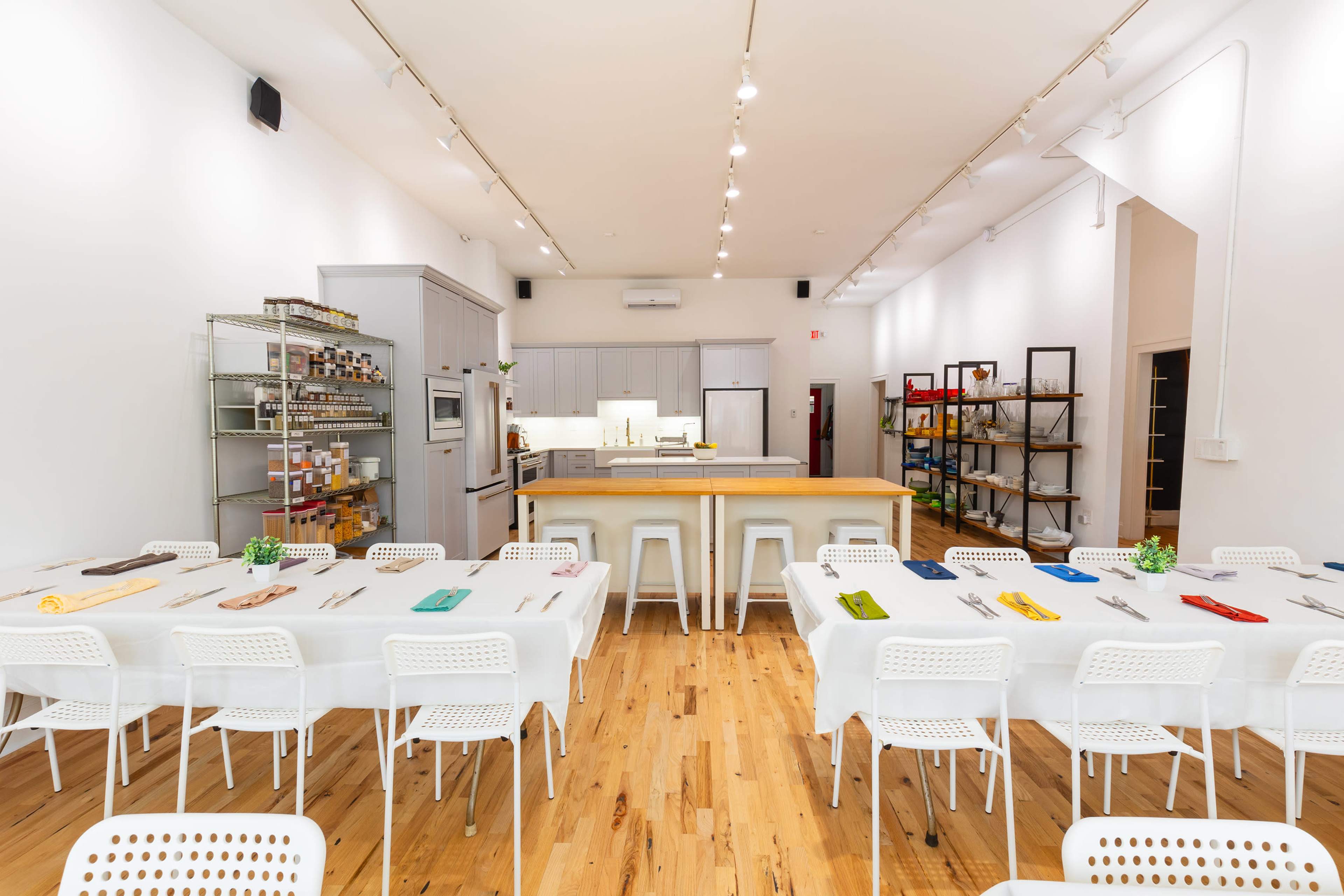 The image shows a clean and modern kitchen space equipped with a central island and several tables set up for cooking classes or events.