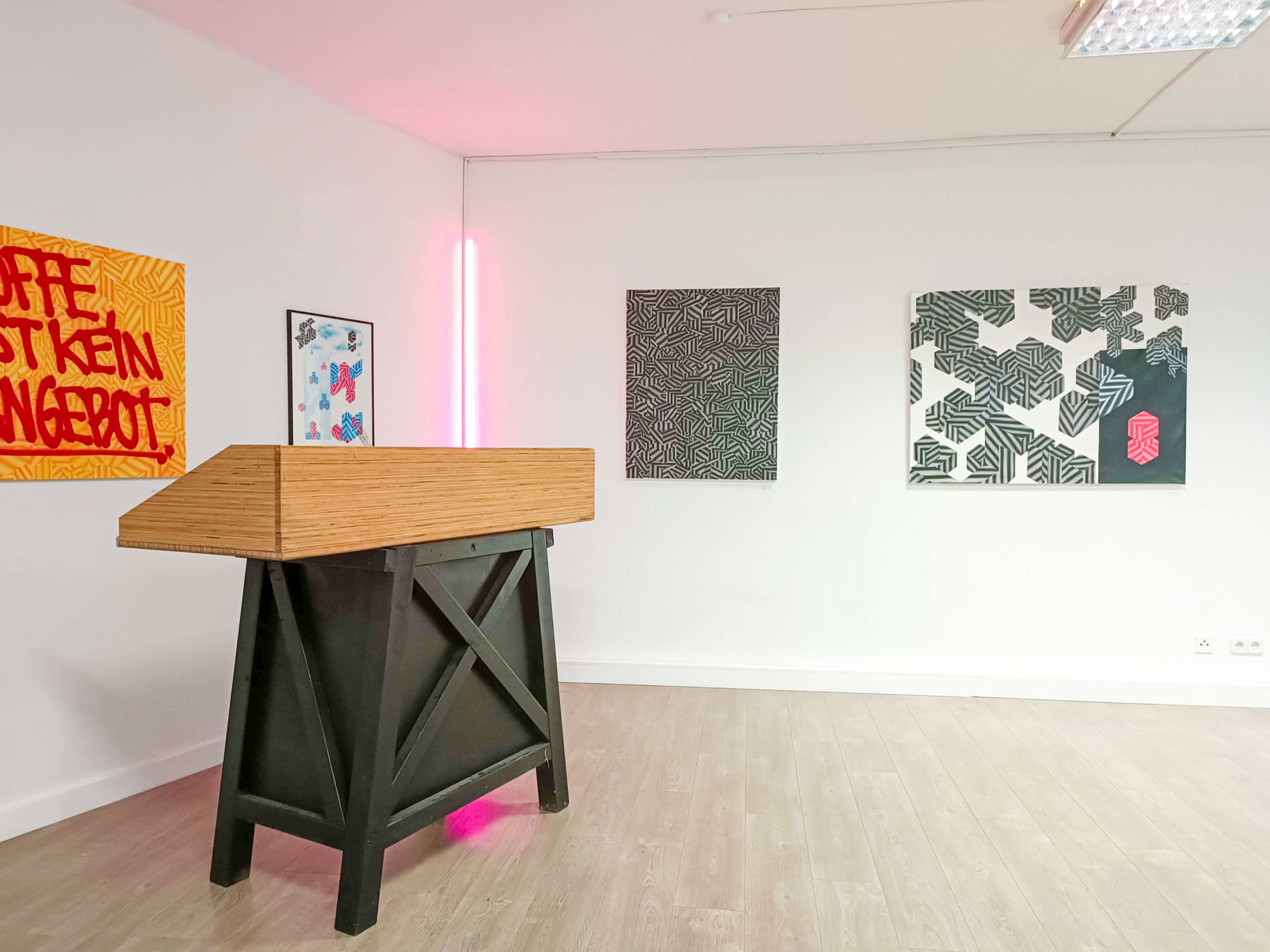 The image shows an art gallery interior with a wooden reception desk and three patterned artworks displayed on the walls.