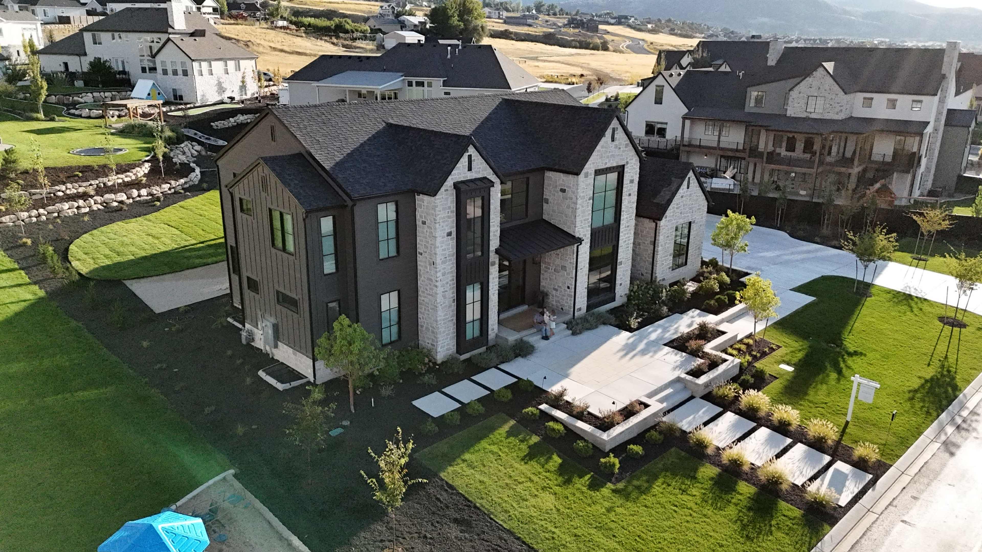 A modern, two-story house featuring a combination of stone and dark wood siding is situated on a landscaped lot with a path and garden beds.