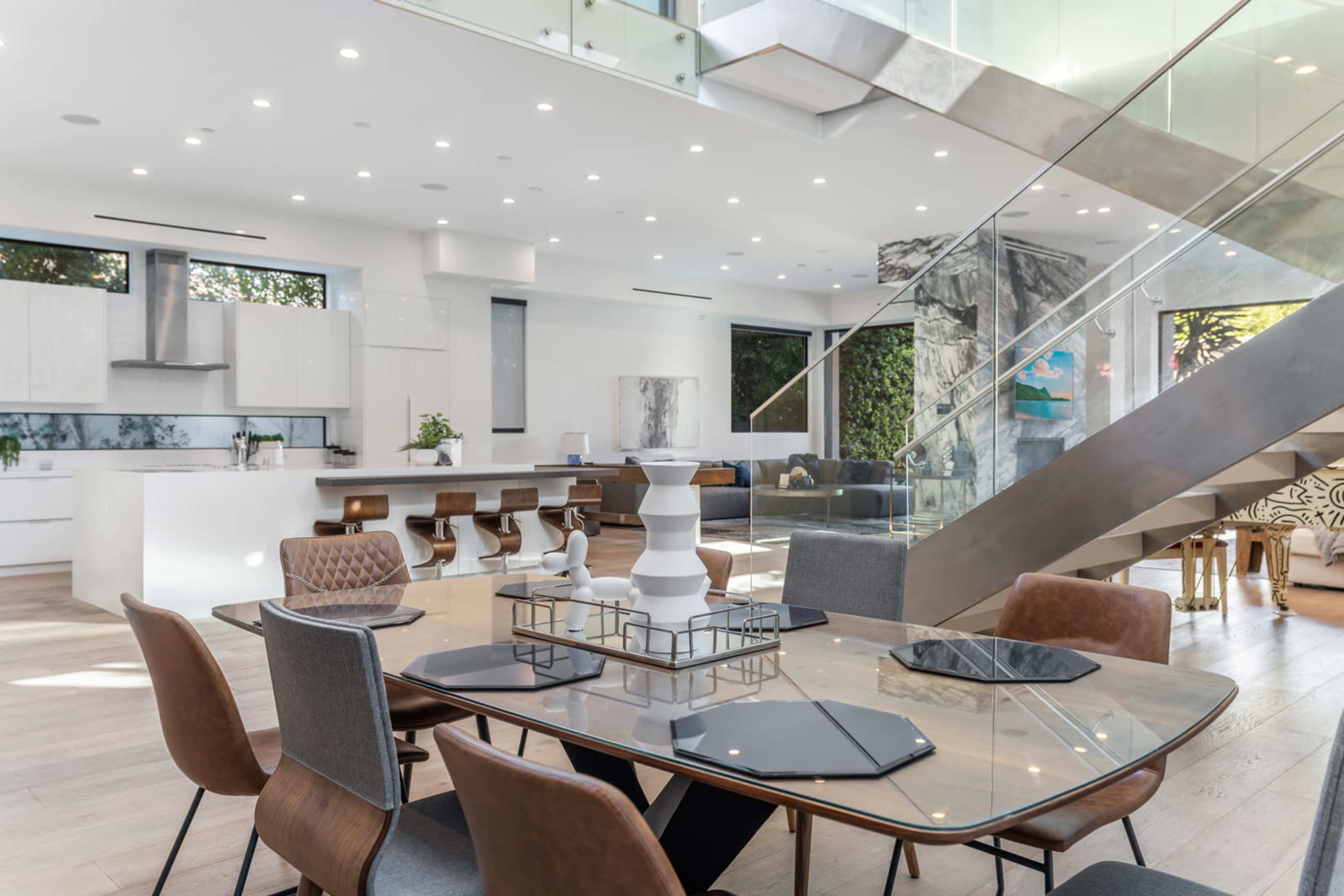 A modern dining area with a glass table surrounded by various chairs, adjacent to a spacious kitchen and a staircase leading to an upper level.