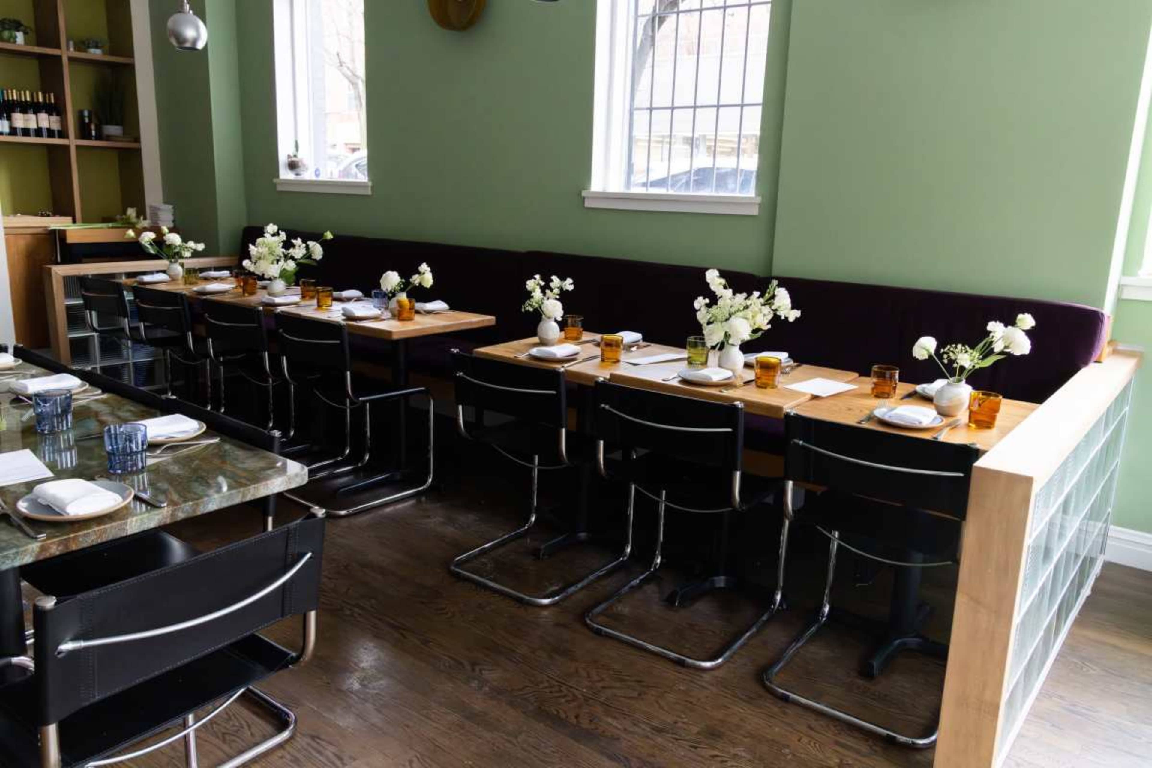 The image shows a restaurant interior with a row of neatly set tables featuring floral arrangements and glassware, against a green wall.