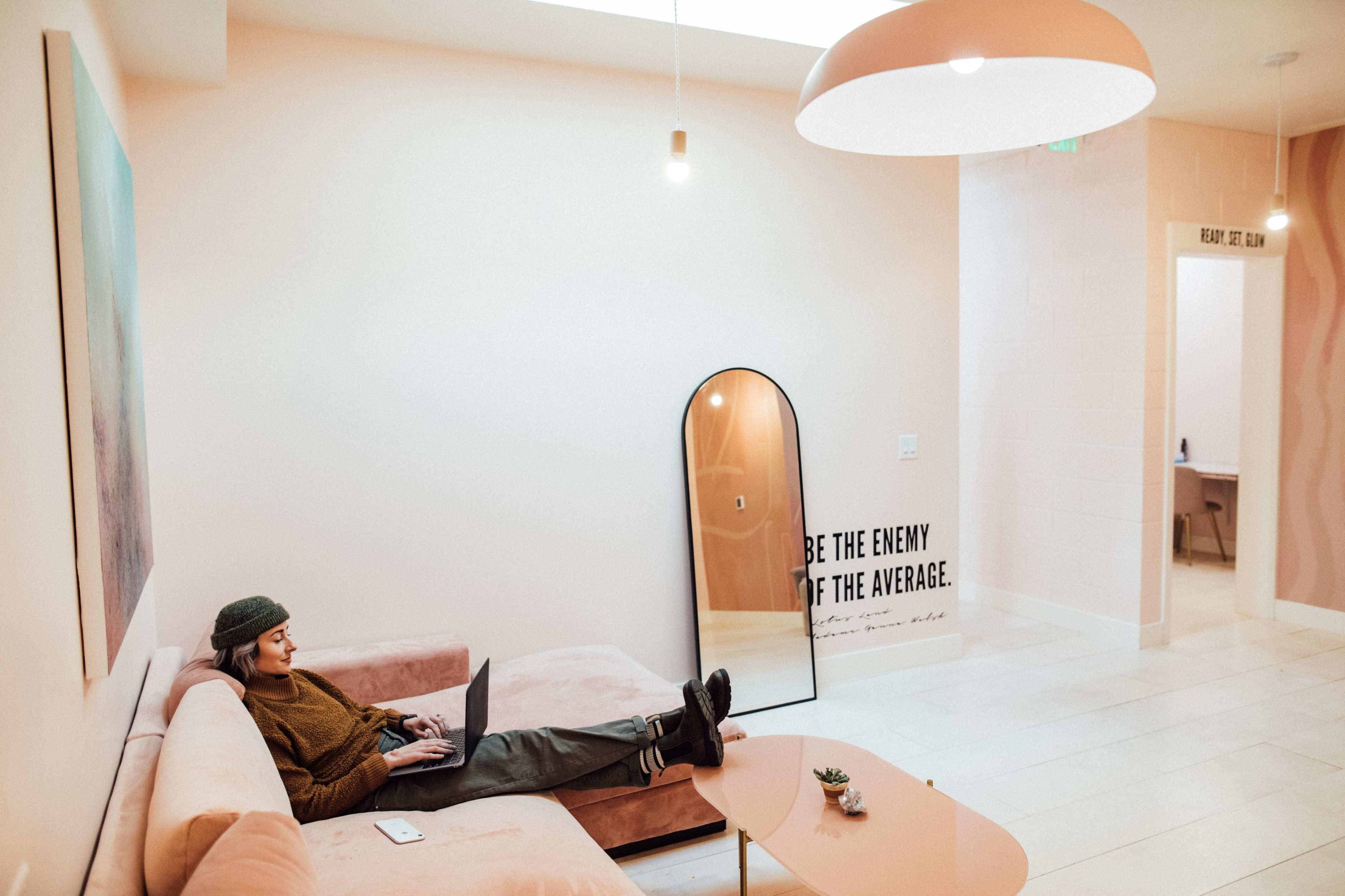 A person sits on a pink couch working on a laptop in a modern, softly lit room with a mirror and a motivational quote on the wall.