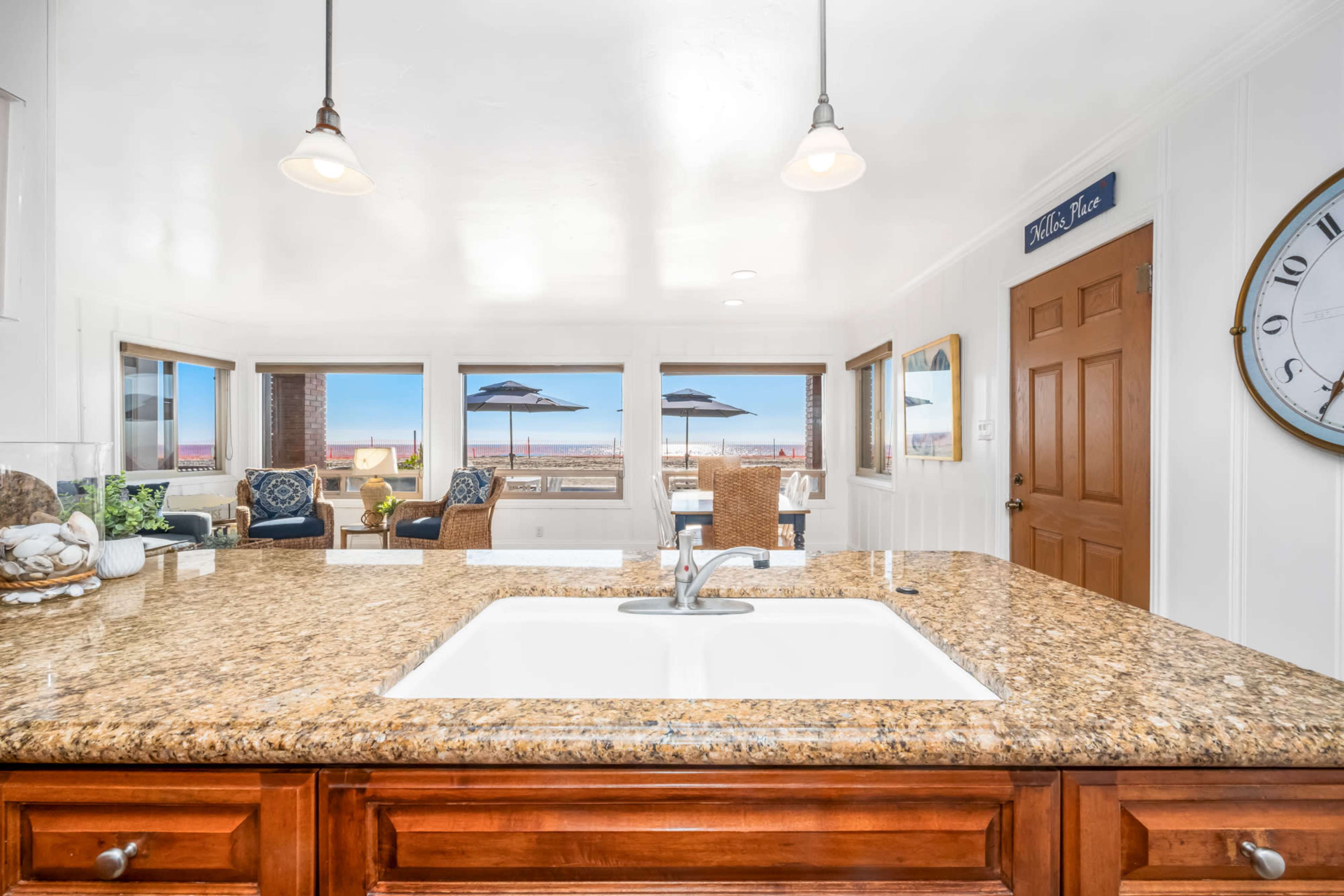 A kitchen with a granite countertop overlooks a living area with seating and a view of the beach through large windows.