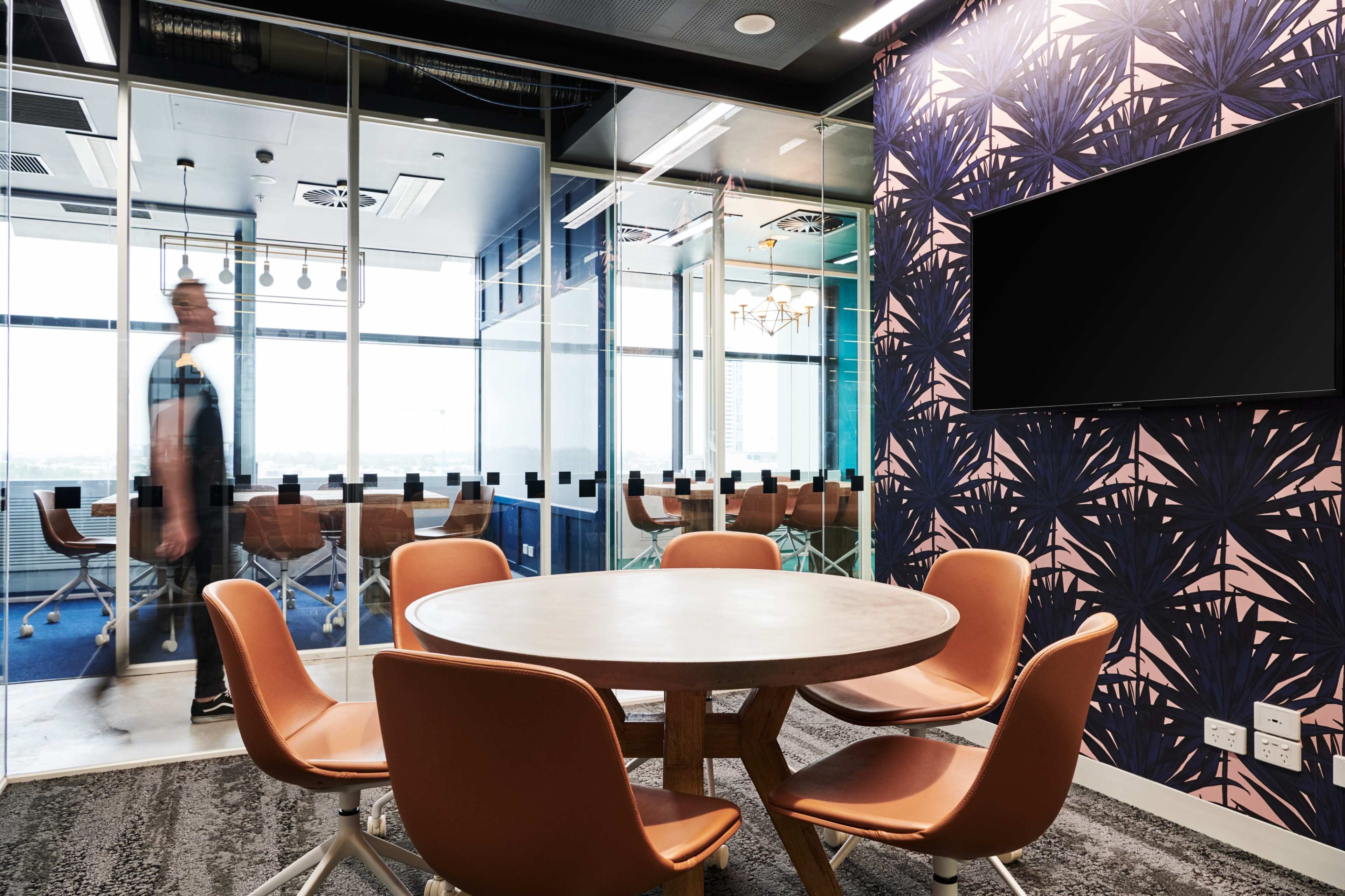 A modern conference room features a circular table with brown chairs, glass walls, and a wall adorned with tropical-patterned wallpaper.