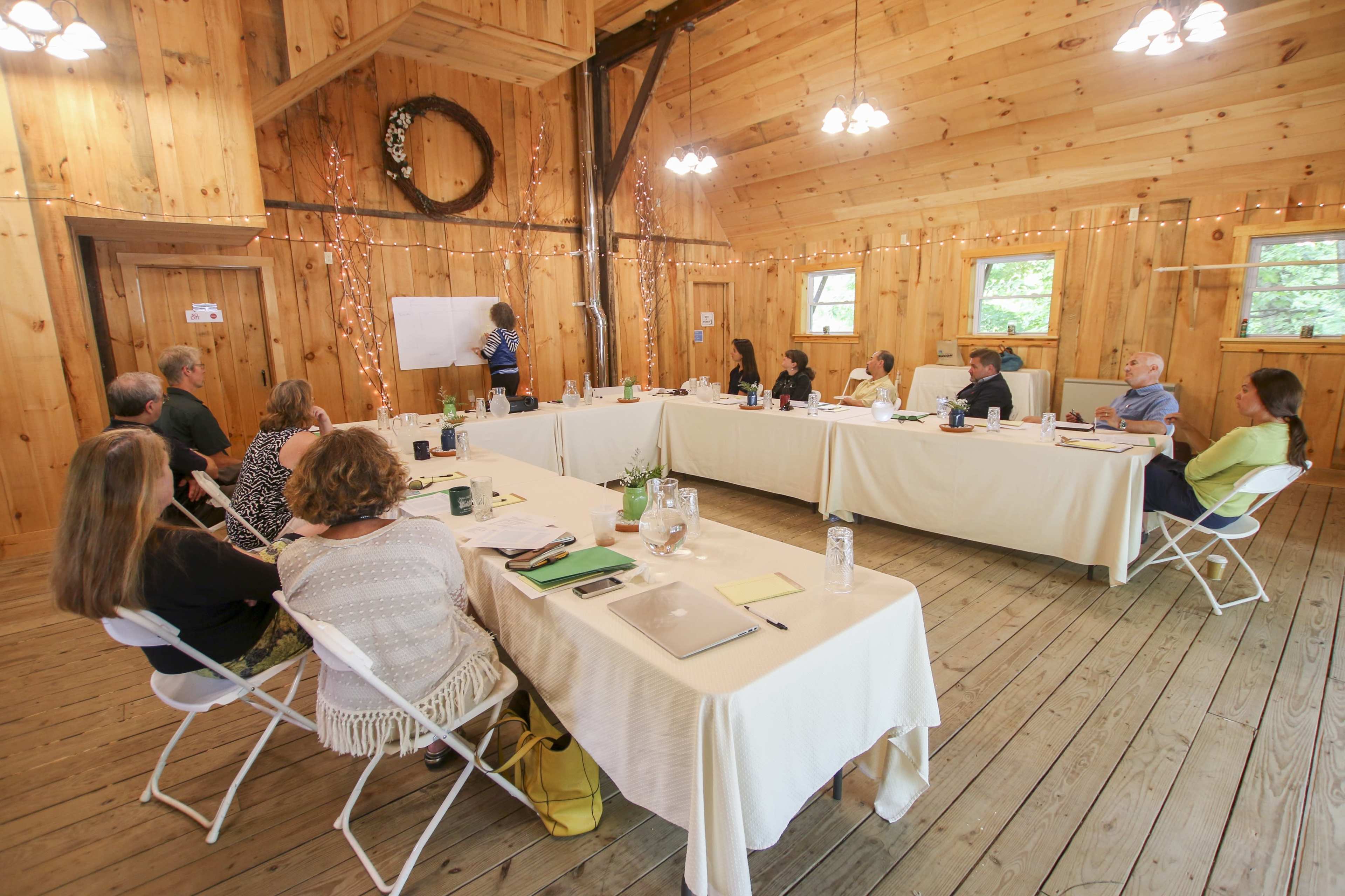 A group of people is seated at tables in a wooden room, while one person stands at a whiteboard giving a presentation.