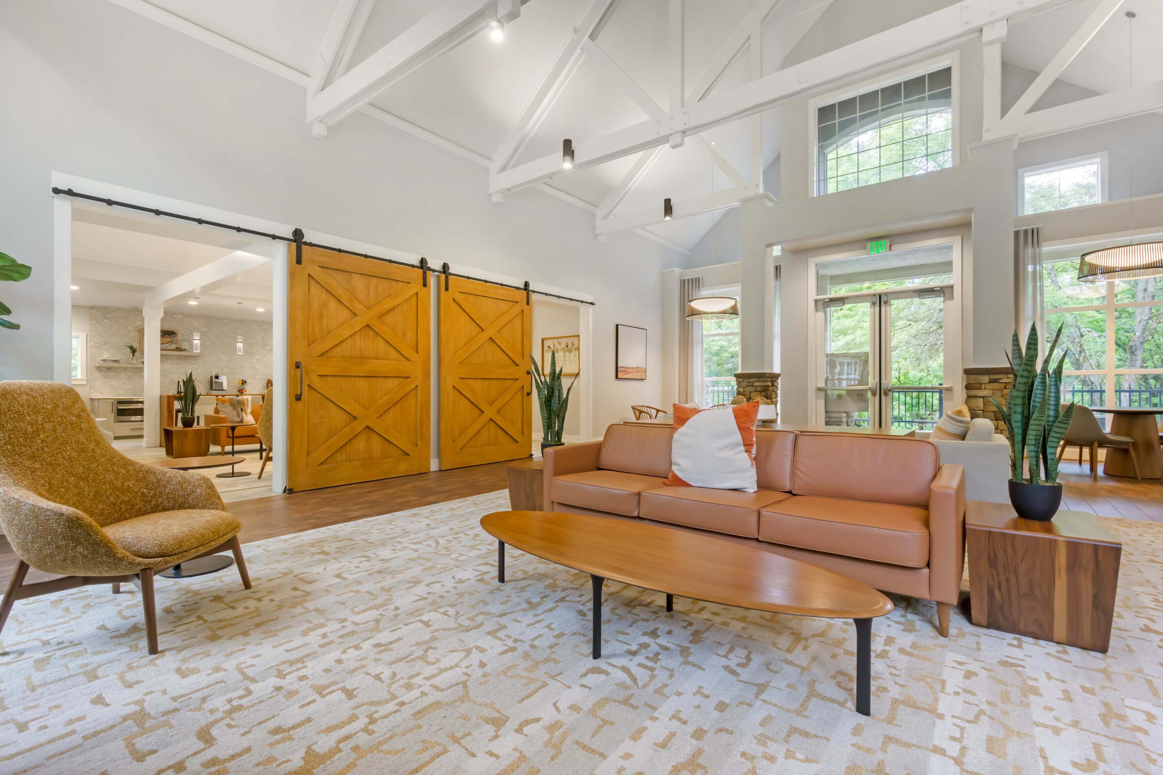 The image shows a modern living space featuring a brown leather sofa, a wooden coffee table, and sliding barn doors, with large windows that provide ample natural light.