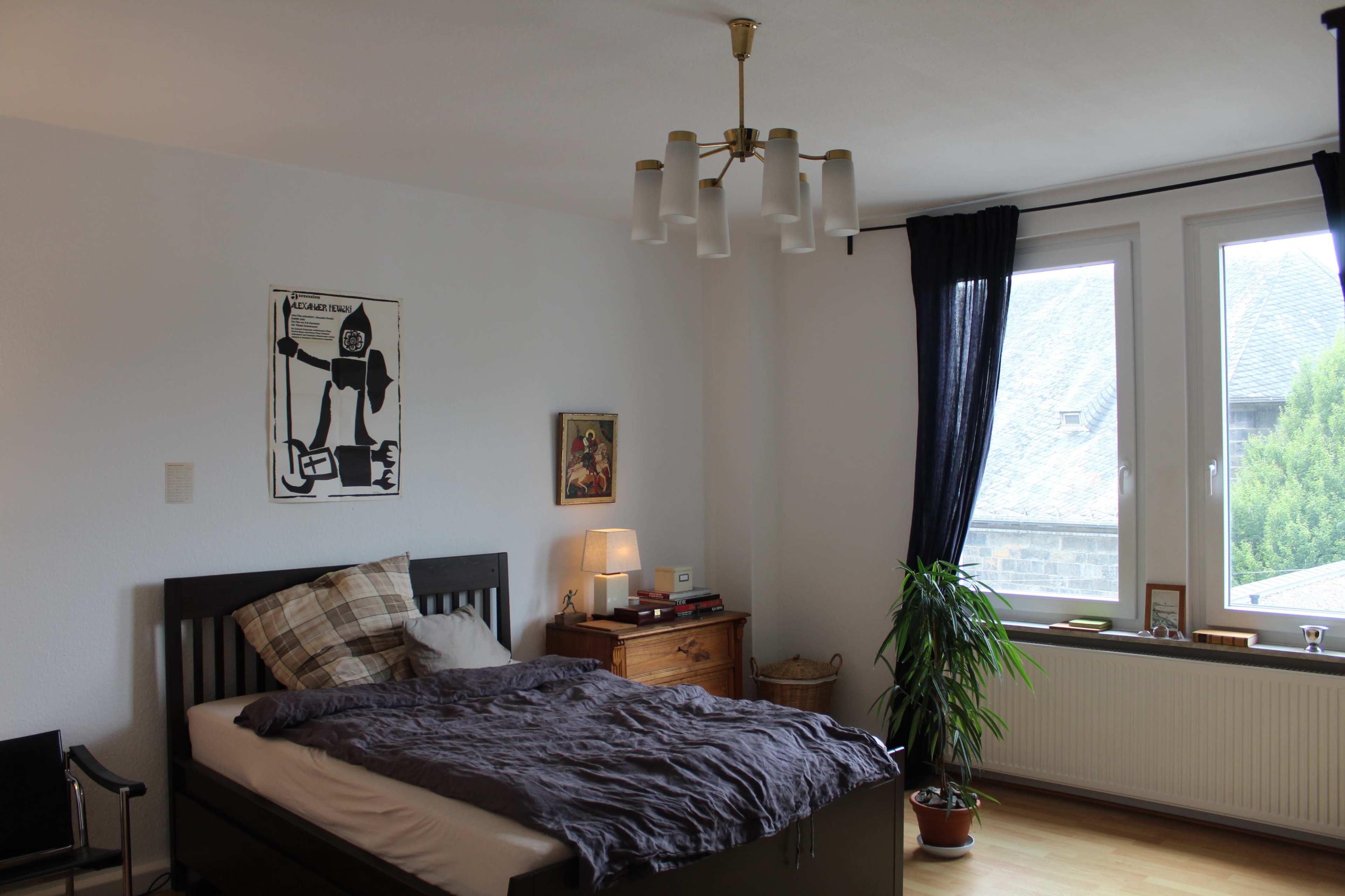The bedroom features a bed with a dark gray duvet, a bedside lamp, a wooden nightstand, a chair, and a large window with dark curtains, illuminated by natural light.