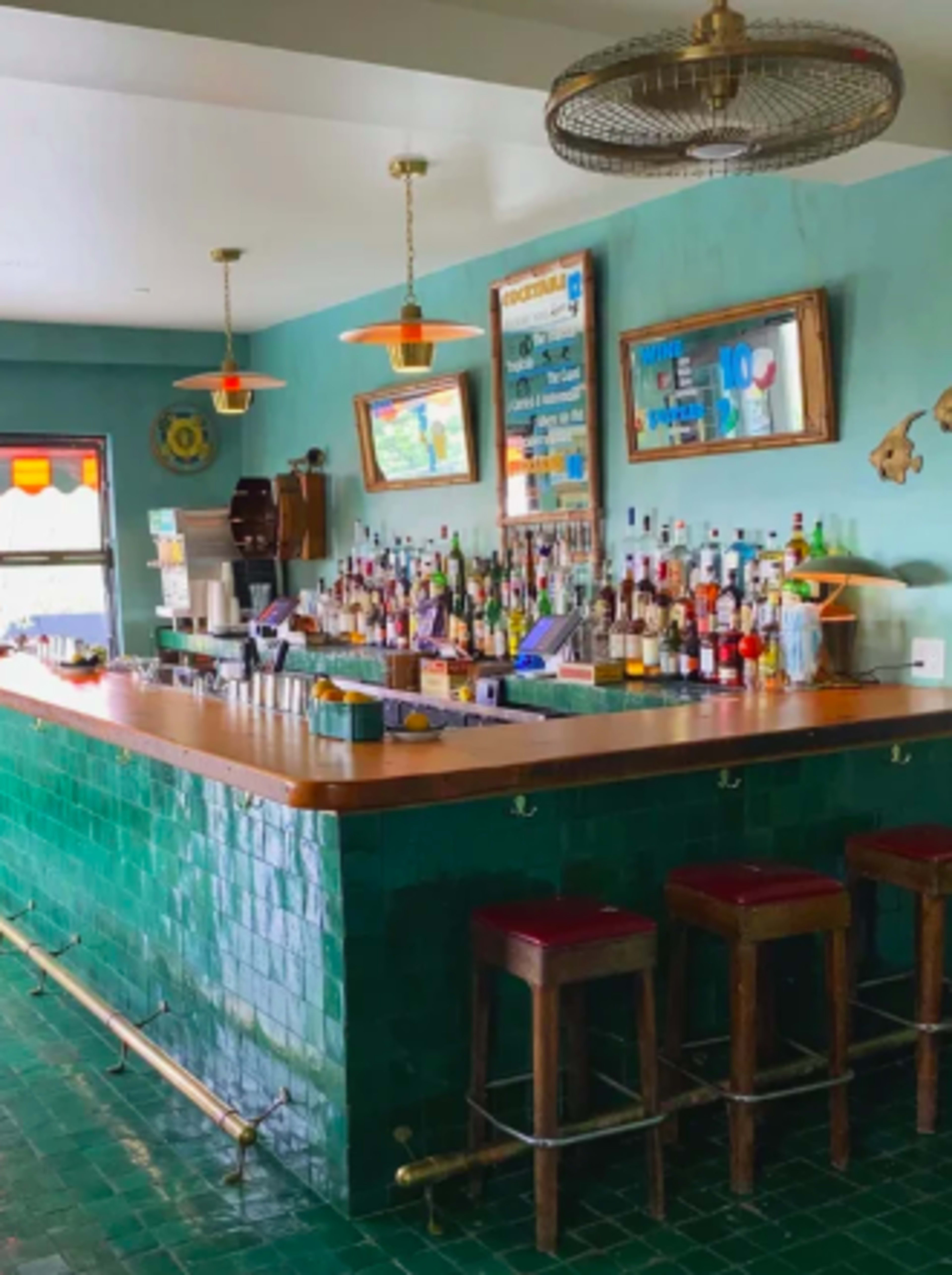 The image shows a brightly colored bar area featuring a green tiled counter, wooden stools, and a variety of liquor bottles displayed behind the bar.